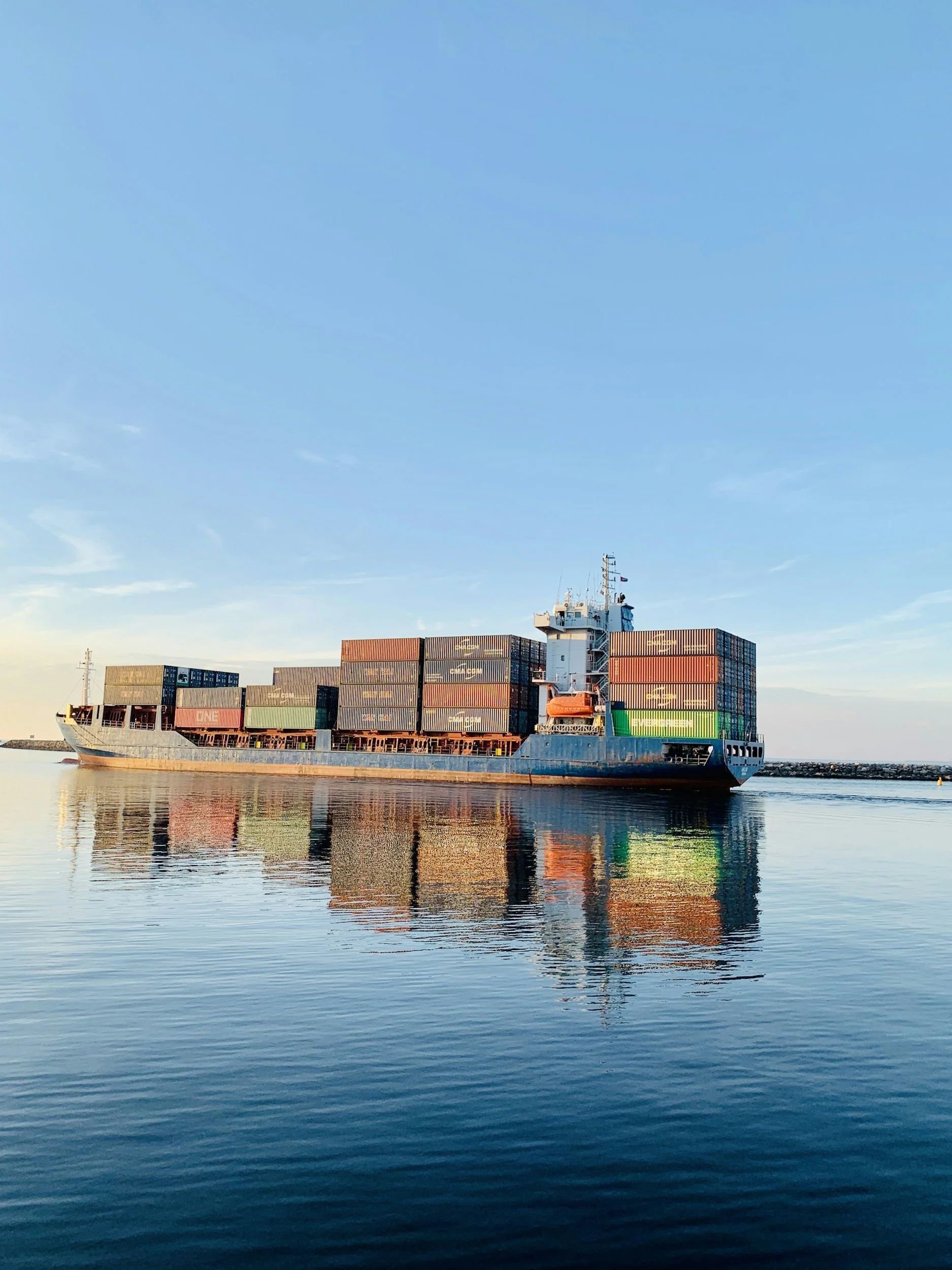 A cargo ship with stacked shipping containers sailing on calm water under a clear blue sky.
