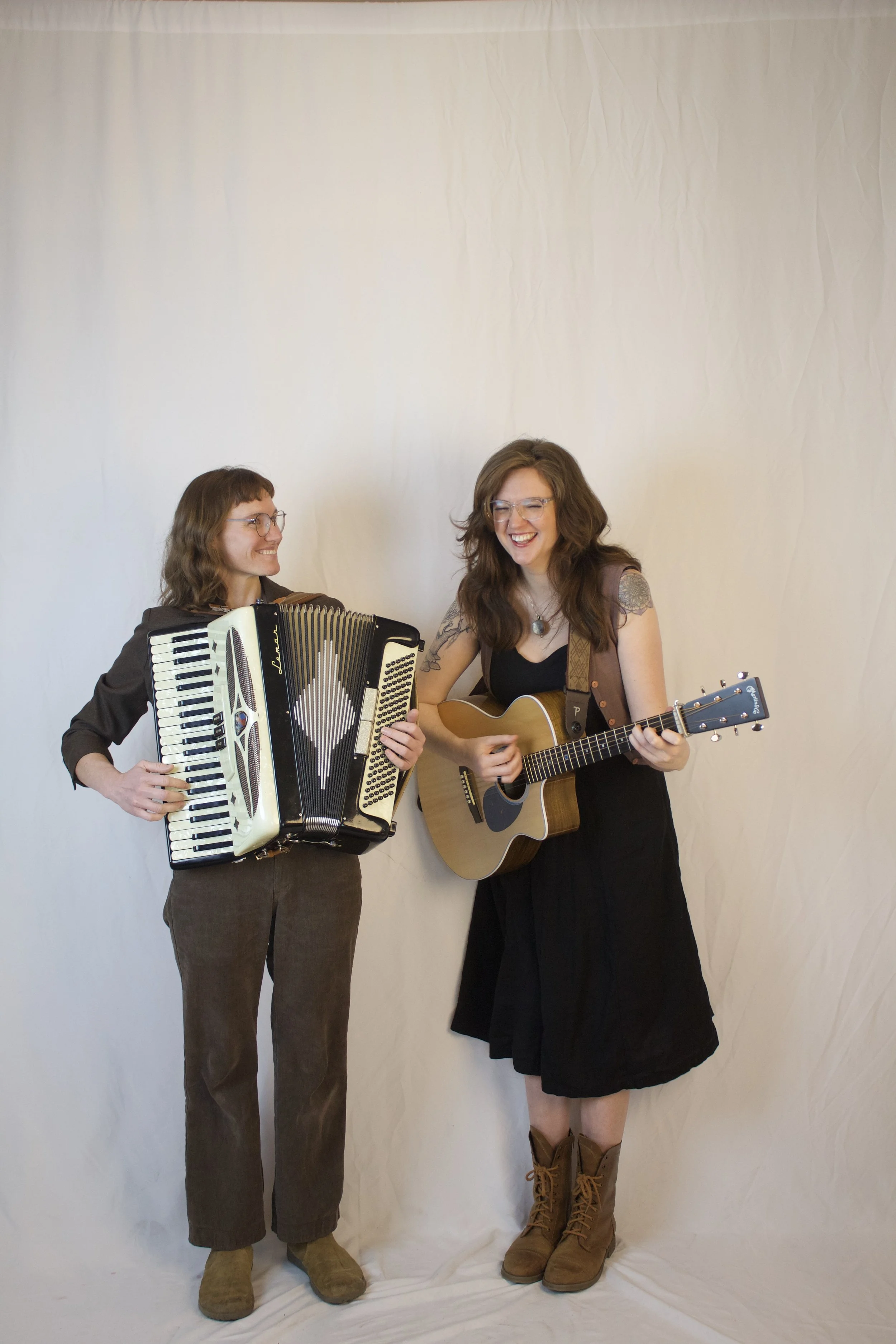 Holler & Crow : Lark Frazier and Ellison Graham playing musical instruments and singing, one with an accordion and the other with an acoustic guitar, standing against a plain white background.