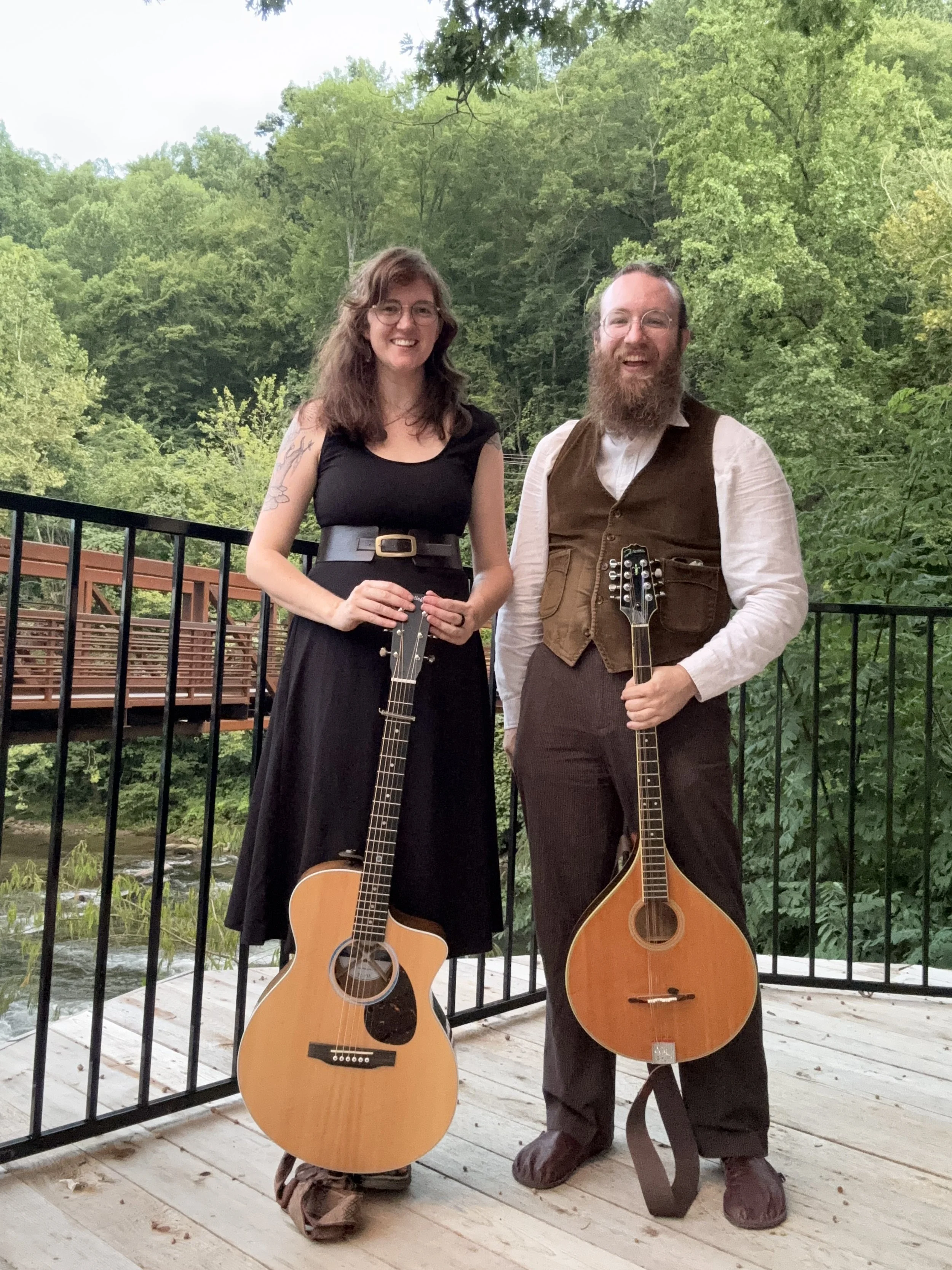 Holler & Crow: Lark Frazier and Jon Snead are standing on a wooden deck outdoors, each holding an acoustic guitar, surrounded by green trees and a river in the background, smiling at the camera.