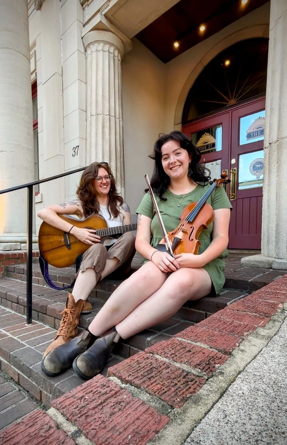 Two women sitting on steps outside a building, one playing guitar and the other holding a violin, both smiling.