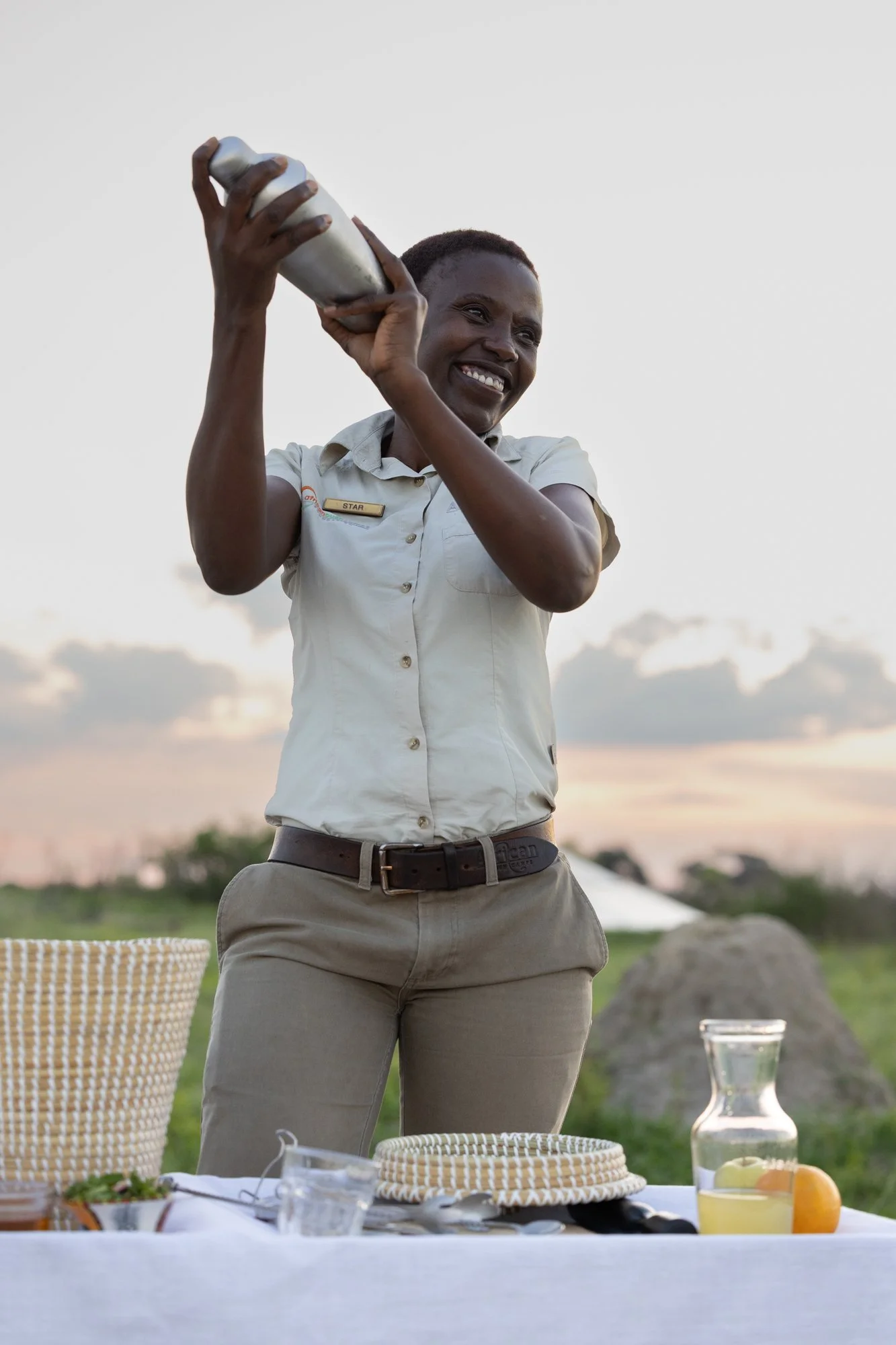 A woman wearing a white short-sleeve shirt with a badge and khaki pants is smiling and making a cocktail in a metal shaker in the outdoors at sunset. Somalisa Camp, African Bush Camps, Hwange National Park, Zimbabwe
