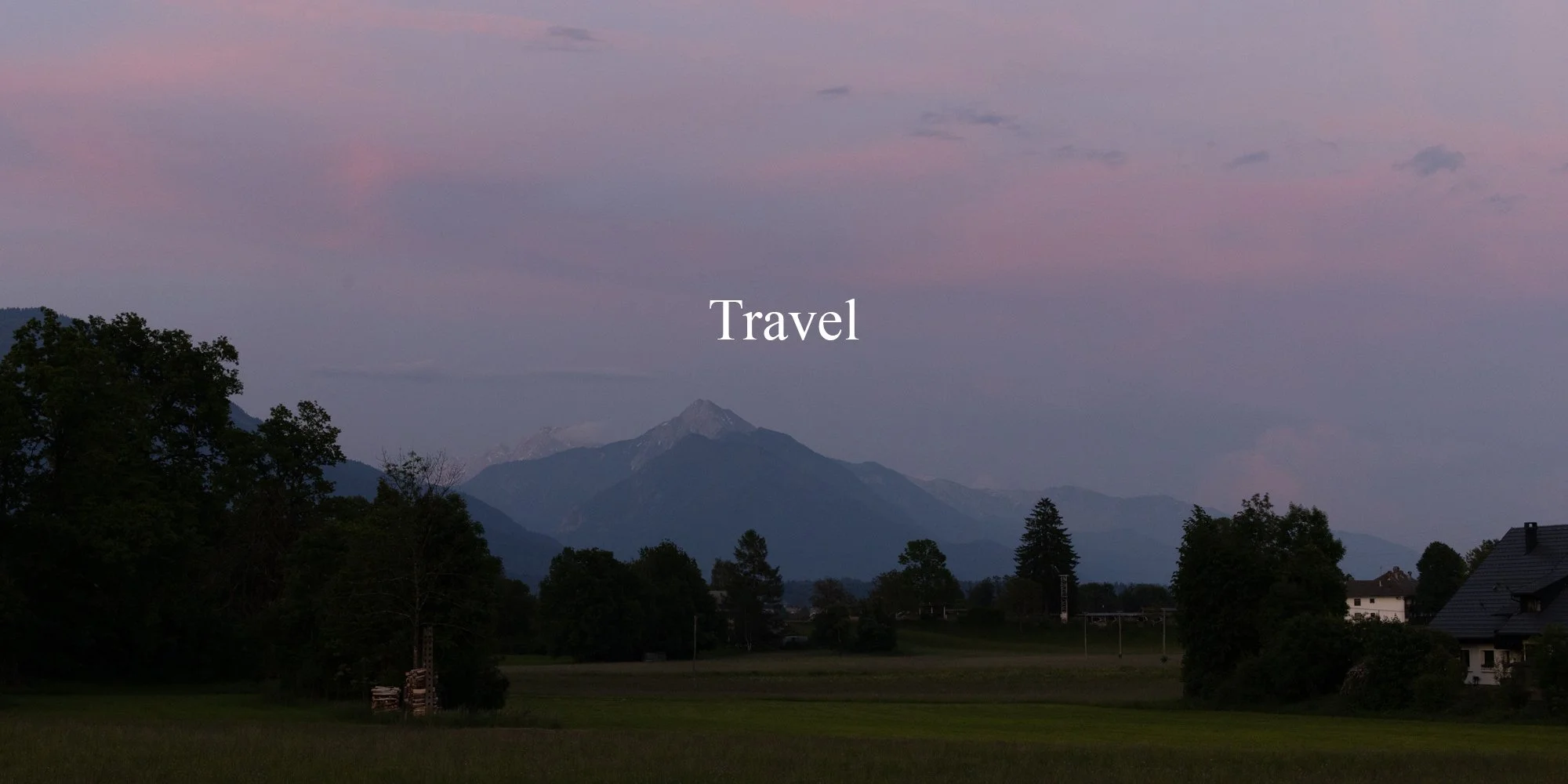A scenic landscape in Bled, Slovenia with tall trees, green fields, and houses in the foreground. In the background, there are mountain peaks under a pastel-colored sky during sunset or dawn with the word 'Travel' centered in white.