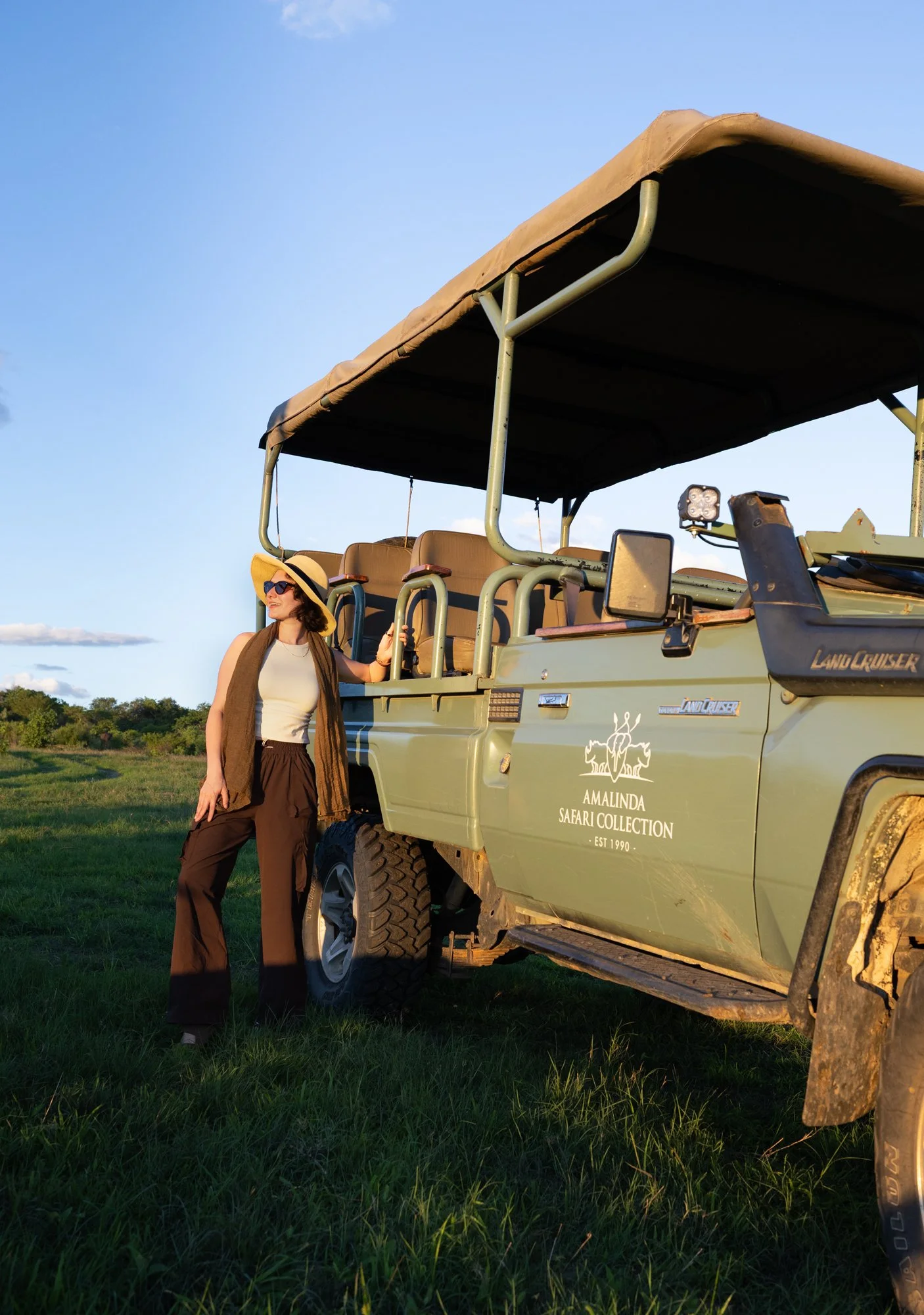 A woman wearing a sun hat, sunglasses, a sleeveless top, and wide-leg pants standing next to a safari vehicle in a grassy field, with a clear blue sky in the background.