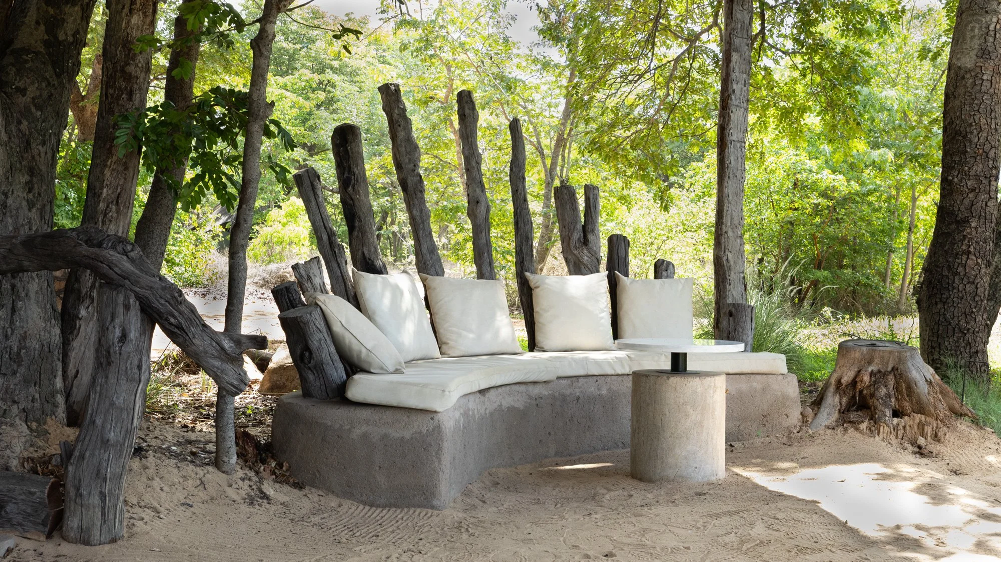 Outdoor seating with white cushions against rustic wooden fence in a forest clearing, sandy ground, and a small round table on a concrete base, surrounded by green trees.