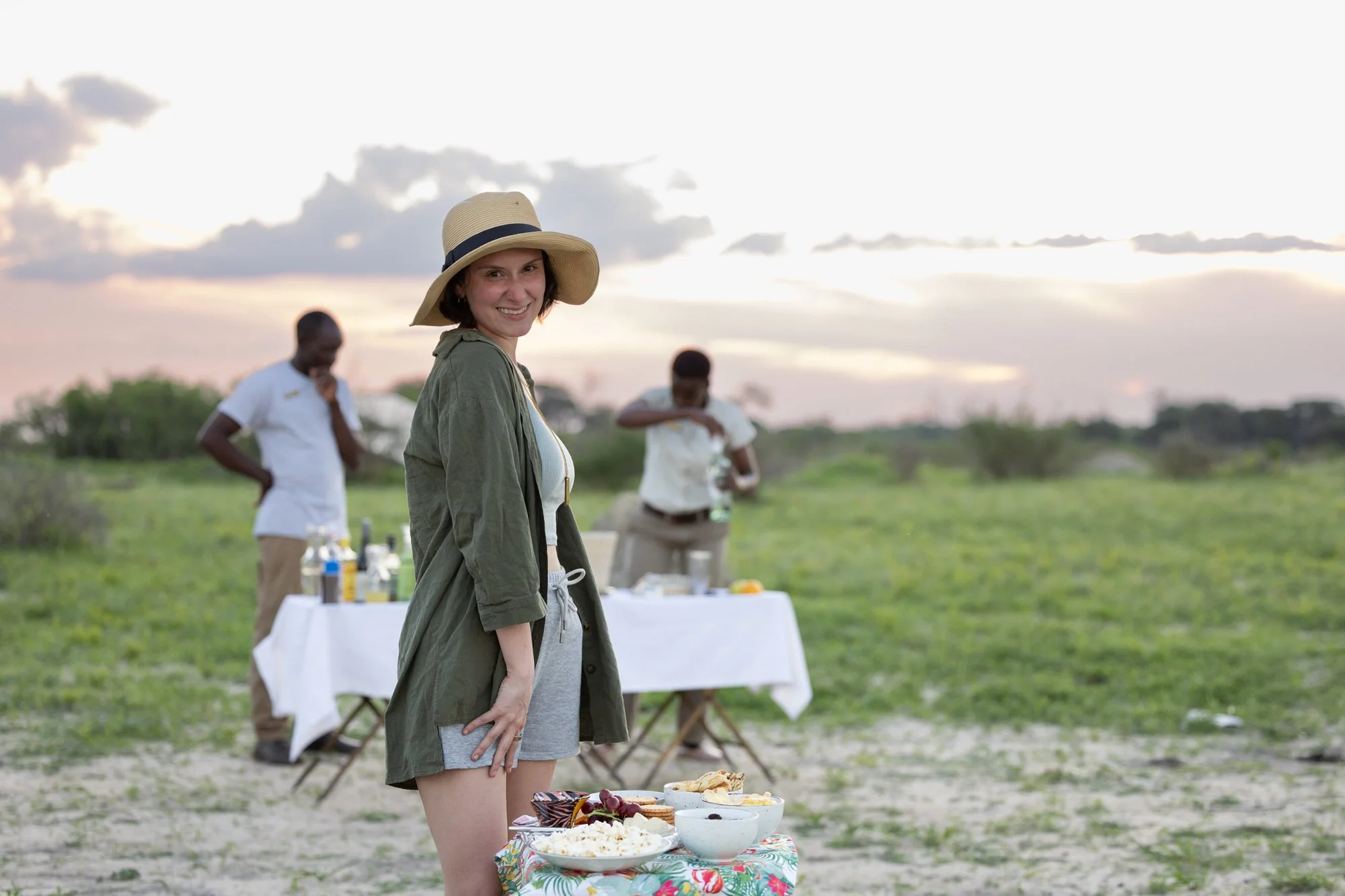 A woman in a wide-brimmed hat smiling at a picnic table with food outdoors during sunset, with two men preparing food or drinks in the background.