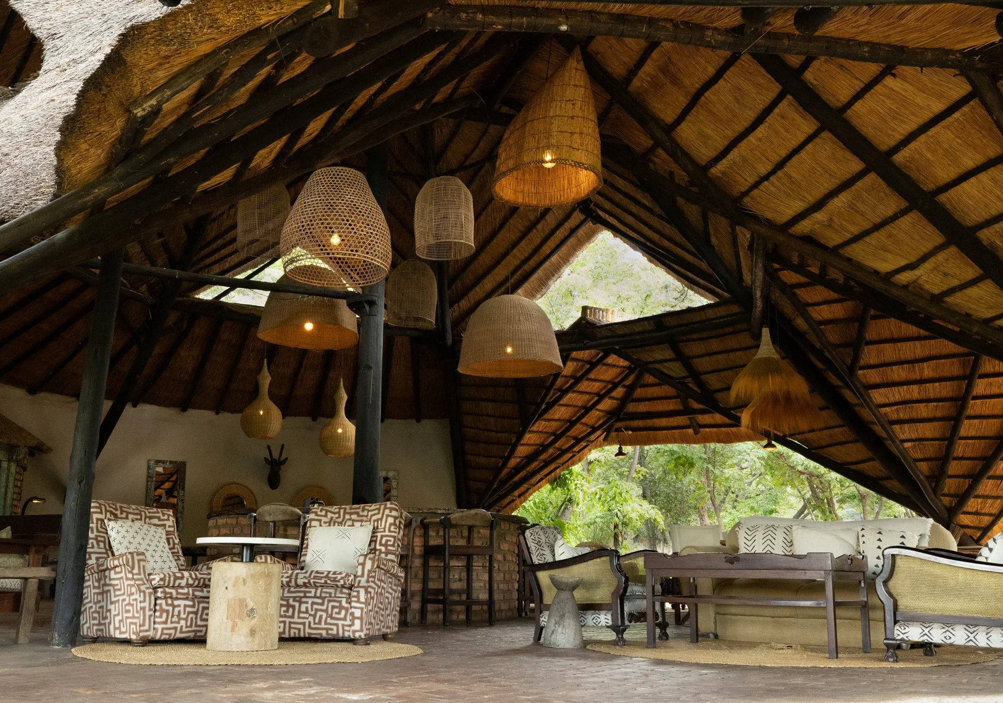 Interior of a rustic open-air lounge with a thatched roof, wicker hanging lamps, and various seating areas with patterned and neutral-colored furniture, surrounded by greenery.