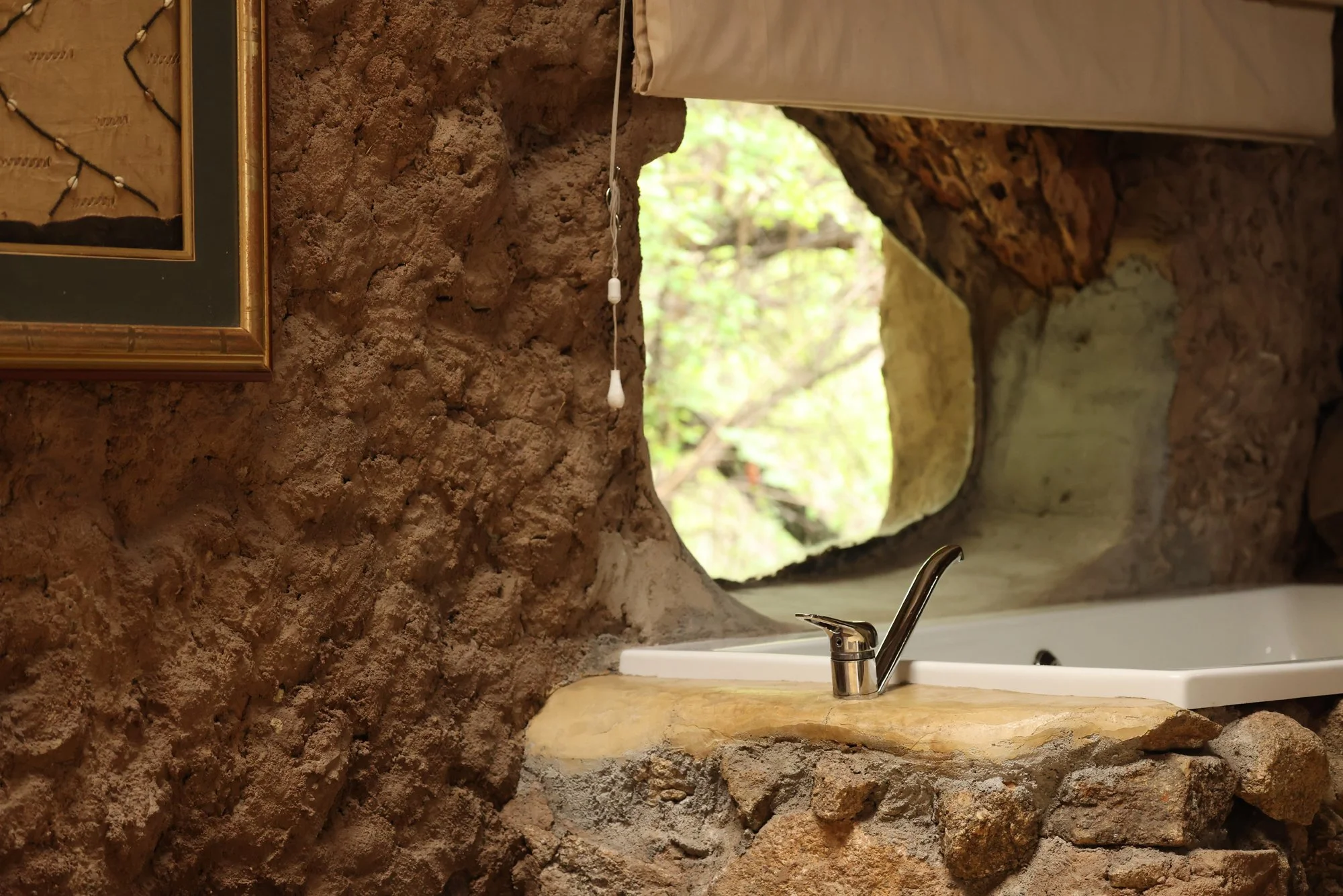 A rustic bathroom with stone walls and a countertop with a small sink and a chrome faucet. There is a round mirror with a natural stone frame. A window in the shape of a cave opening shows trees and greenery outside.