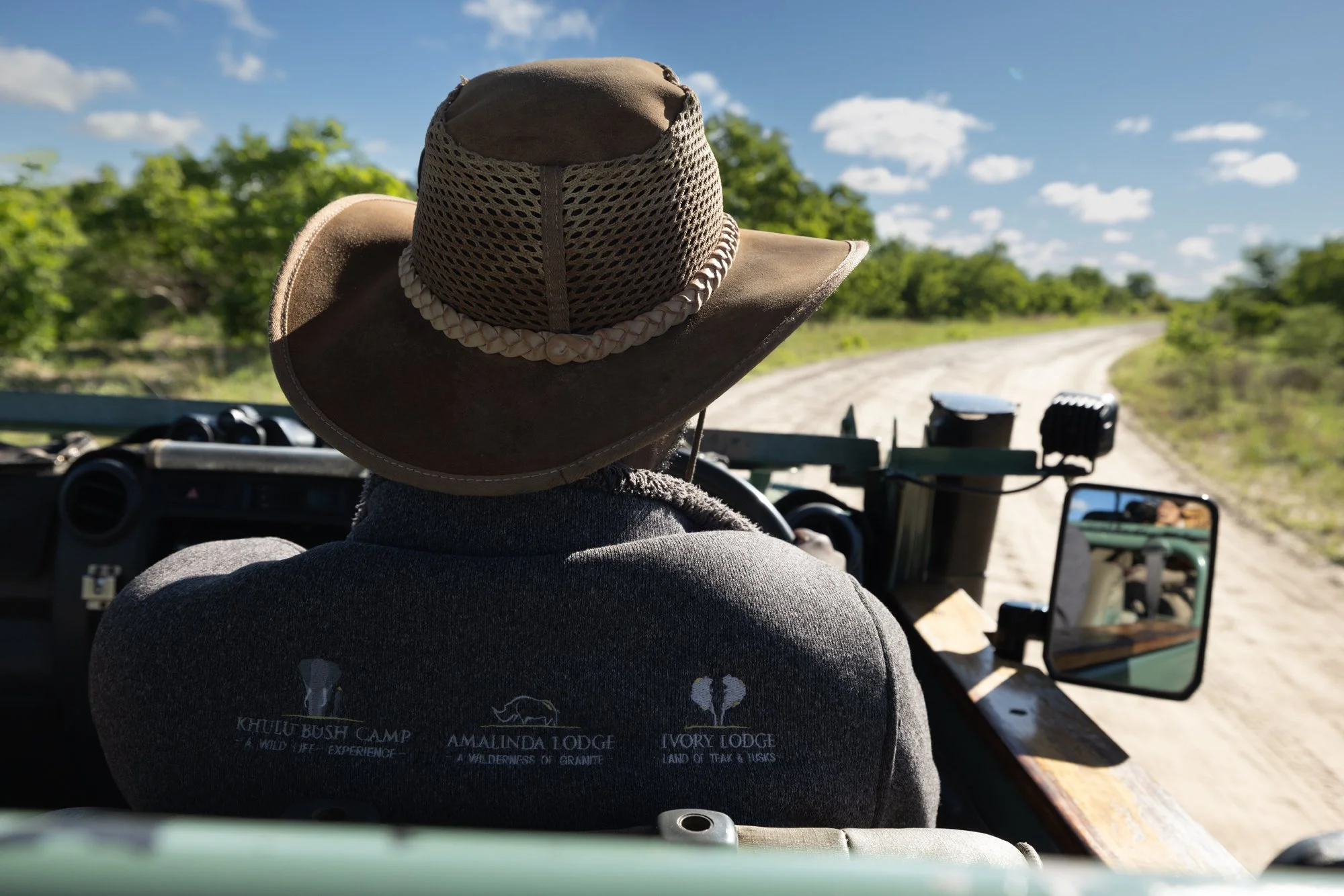 Person wearing a wide-brimmed hat and a dark jacket driving a safari vehicle on a dirt road through a green, wooded landscape with a partly cloudy sky.