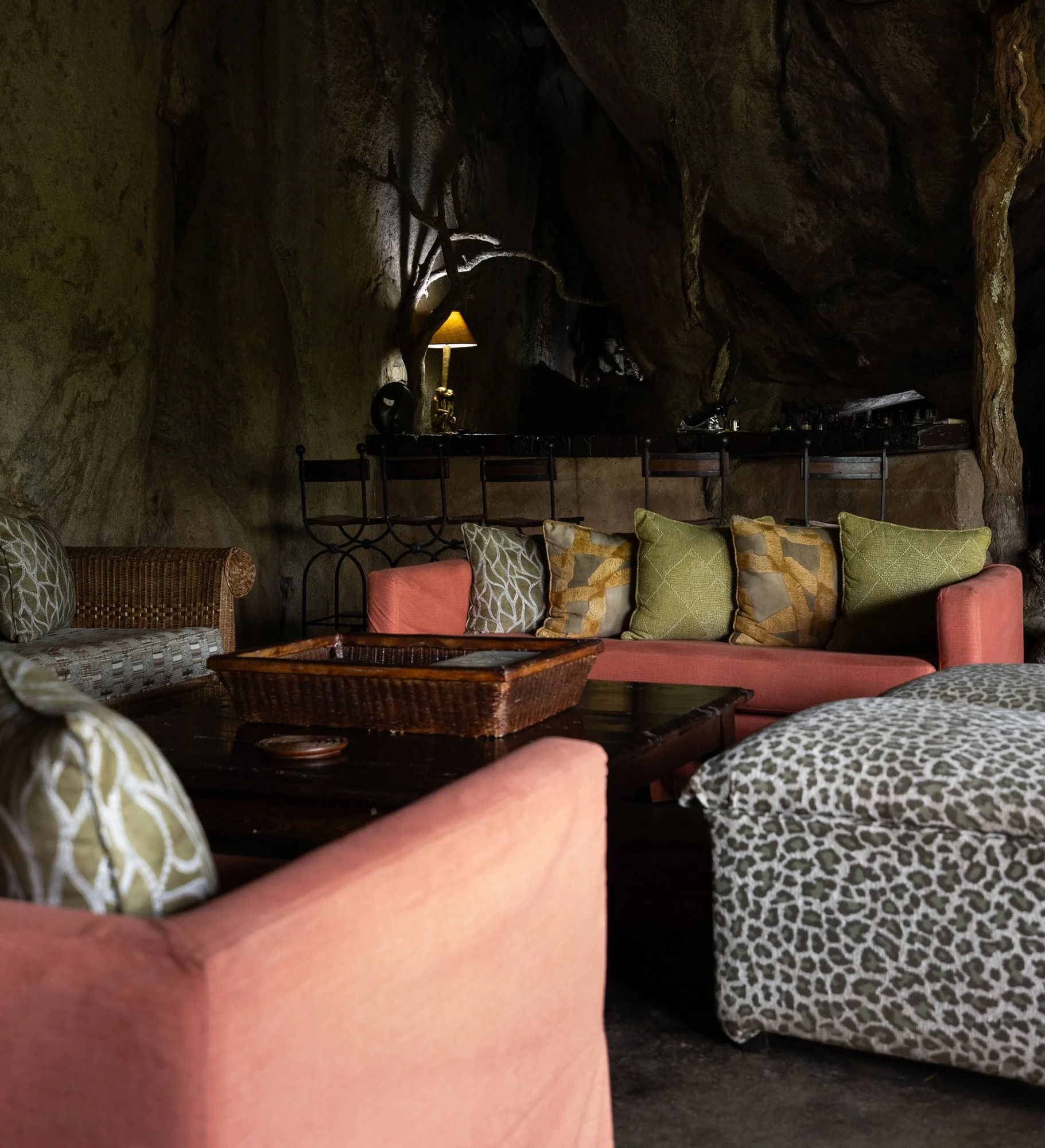 A cozy living room with pink and animal print upholstered furniture, an array of patterned throw pillows, a dark wooden coffee table with a wicker tray, and a rock wall with an embedded lamp in a cave-like setting.