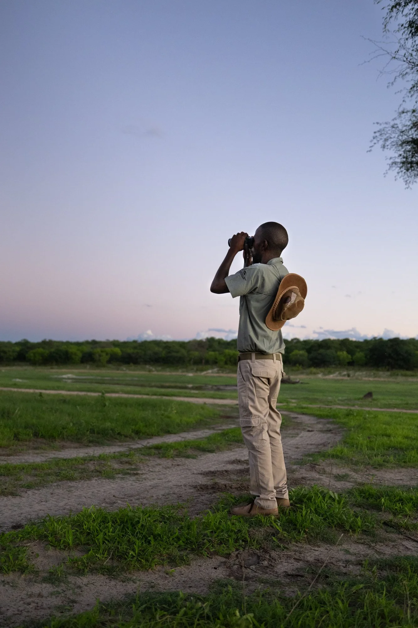 A young man in beige cargo pants and a light-colored shirt, with a hat hanging on his back, standing on a grassy dirt path, looking through binoculars at the horizon during sunset in a rural landscape.