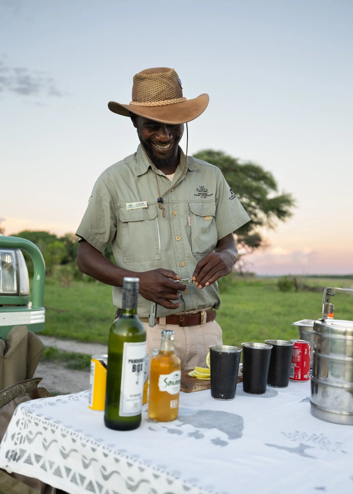 A man in safari attire stands outdoors at sunset, smiling, with a table of drinks including bottles, cups, and cans.