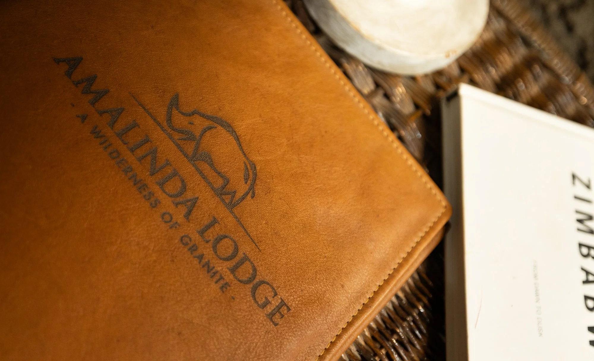 A close-up of a brown leather menu cover with the logo and name 'Amalinda Lodge' embossed on it, placed on a woven surface, with part of a white plate and white sign with black text nearby.