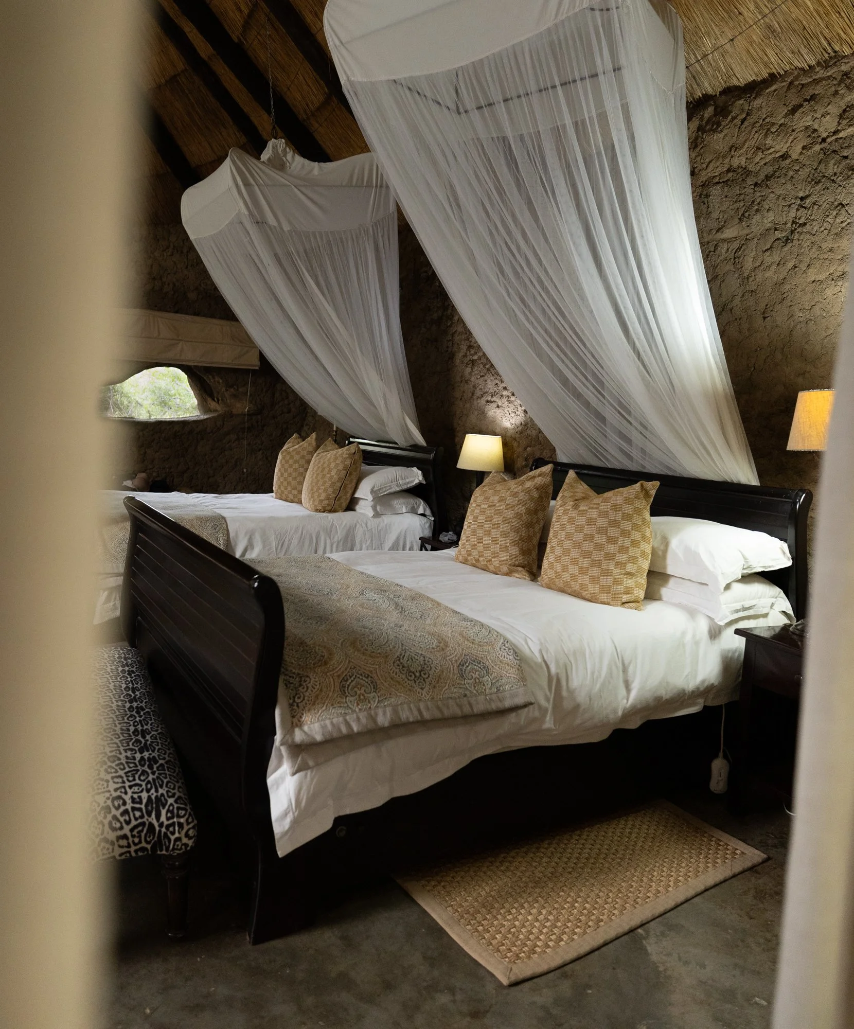 Two beds with mosquito nets hanging from the ceiling in a rustic room with stone walls, wooden ceiling, and lamps. Amalinda Lodge, Matobo National Park, Zimbabwe