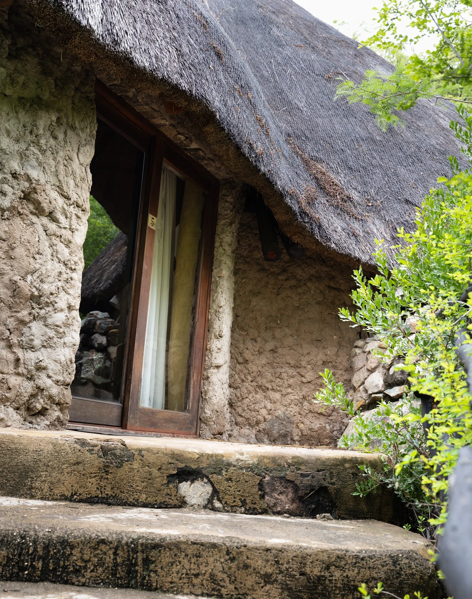 Close-up of a rustic stone and thatch cottage with a sliding glass door, stone steps, and greenery surrounding it.