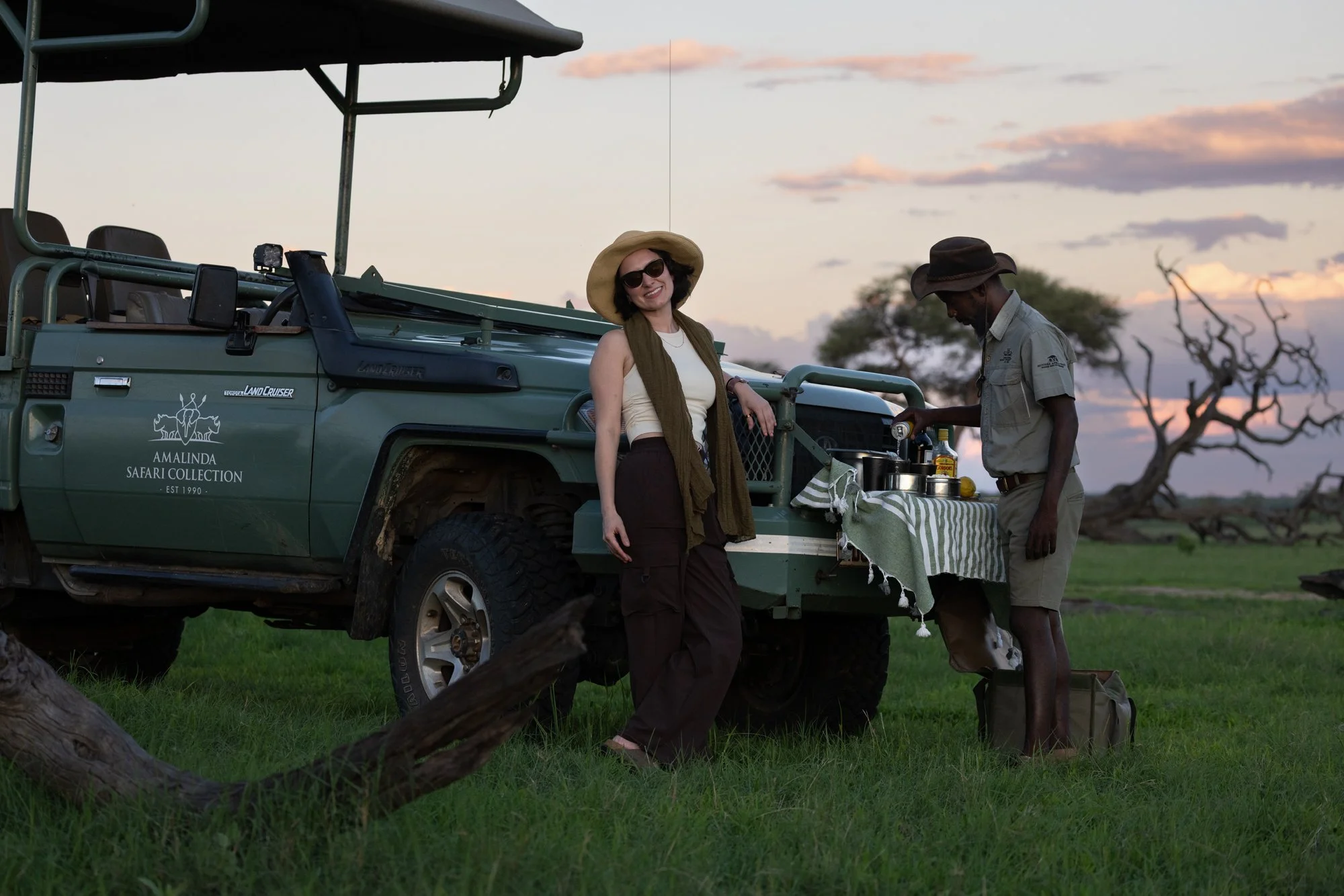 A woman in safari attire standing next to a man preparing drinks at a mobile outdoor bar on the back of a green safari vehicle during sunset in a grassy landscape with a leafless tree in the background.
