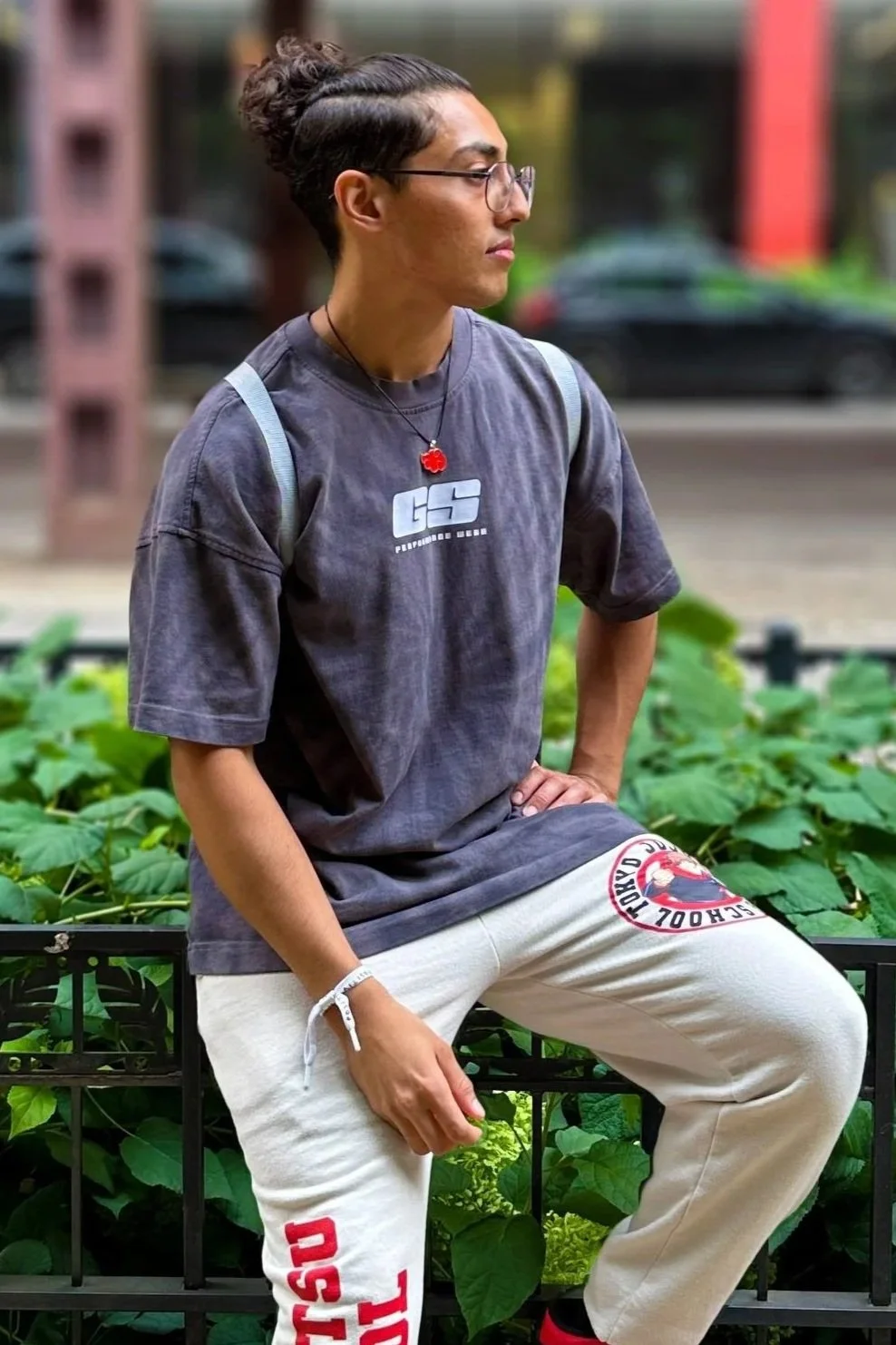A young man with glasses and a bun hairstyle, wearing a dark gray t-shirt with a logo and white sweatpants with red lettering, sitting on a black metal bench in a green outdoor area.