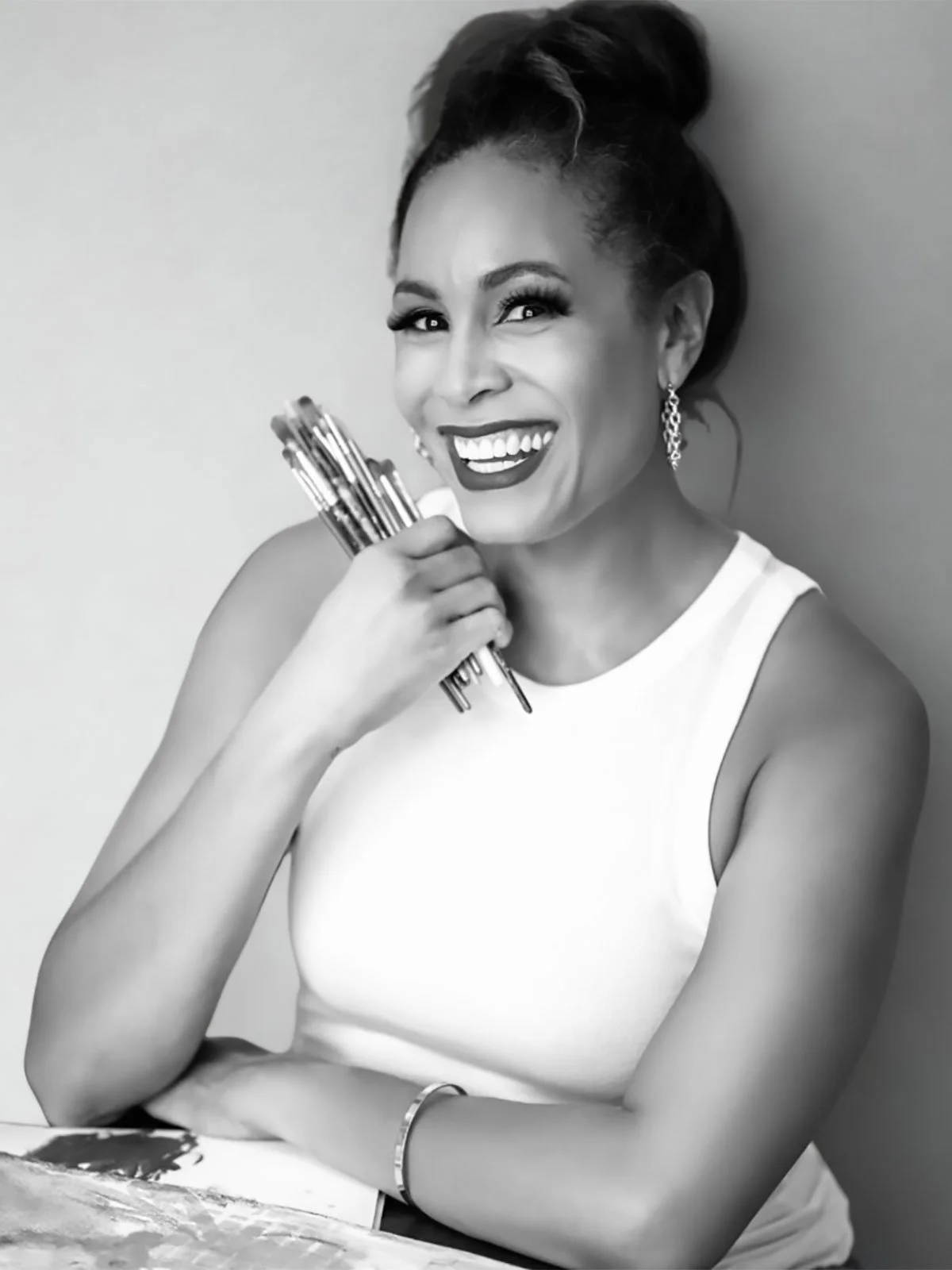 Woman wearing a white tank top and holding paint brushes smiling