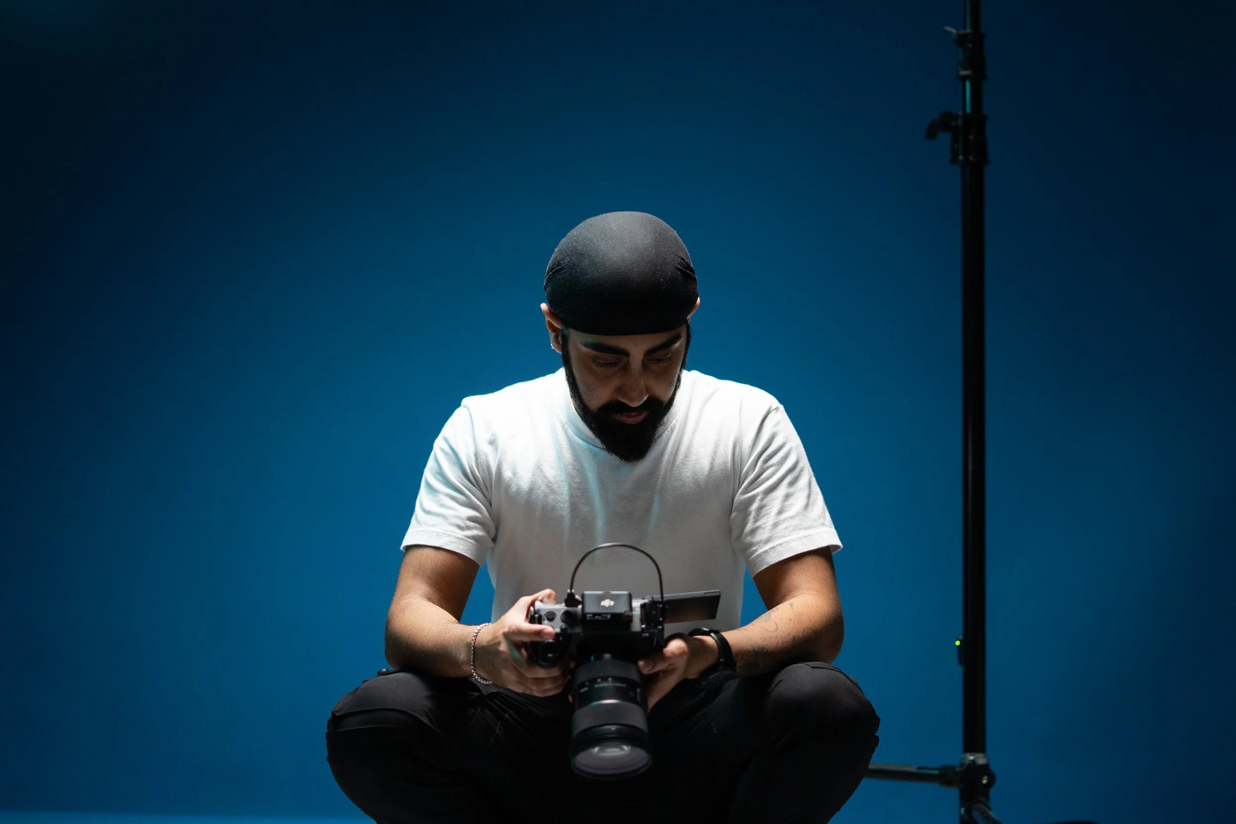 A man wearing a black beanie and white t-shirt looking down at a camera he is holding, with photography equipment and a light stand in the background, against a blue wall.