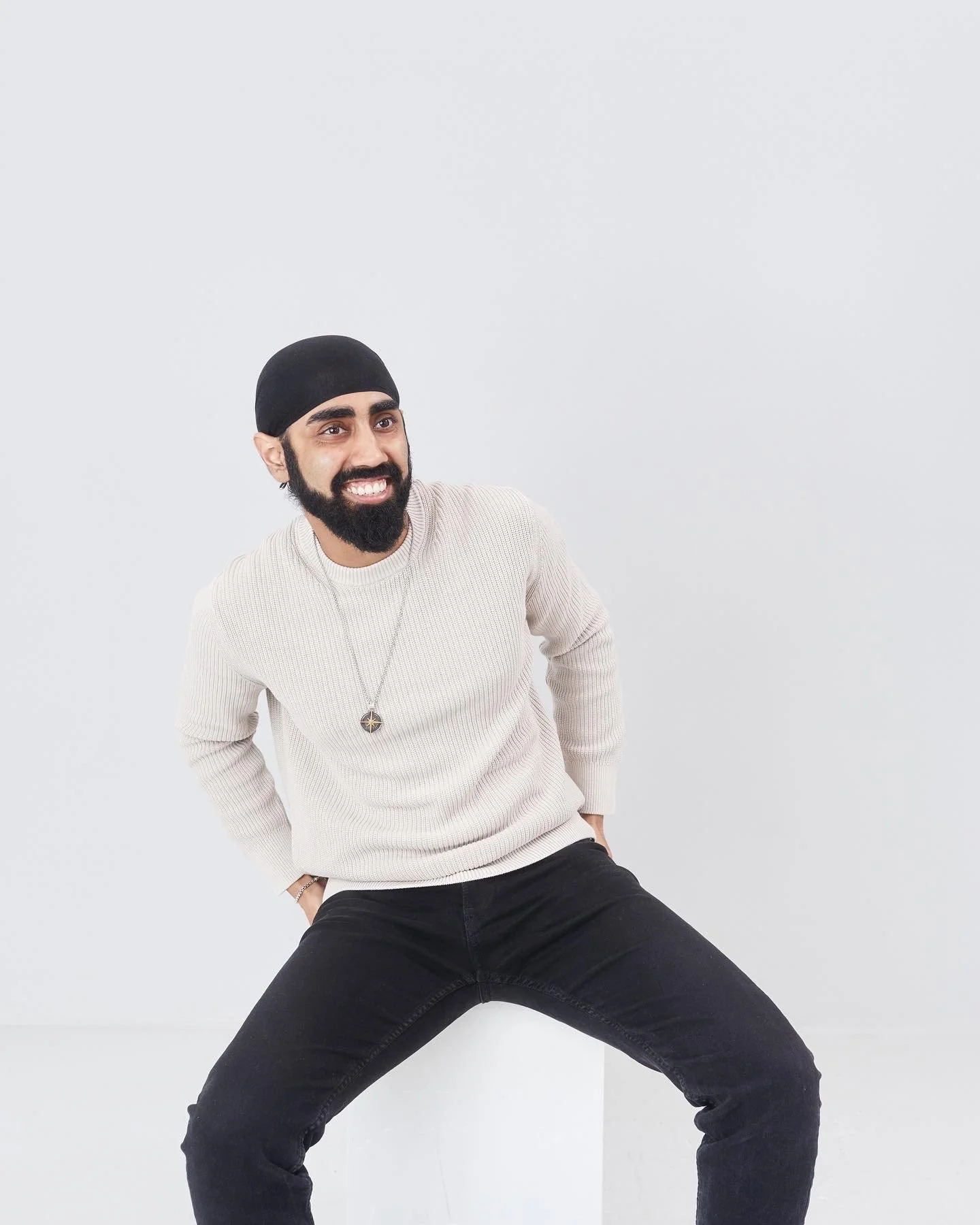 Man with a beard and mustache wearing a black turban, beige sweater, black pants, smiling, posed with legs wide apart and hands behind his back against a plain white background.