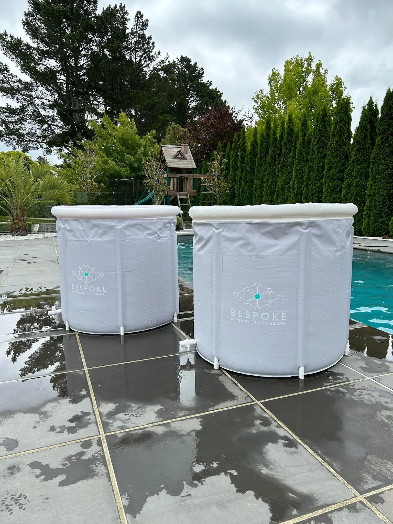 Two white portable hot tubs with the logo 'Bespoke Physiotherapy & Health' on the side placed on a wet tiled pool deck outdoors, with a swimming pool, green trees, a wooden playhouse, and a cloudy sky in the background.