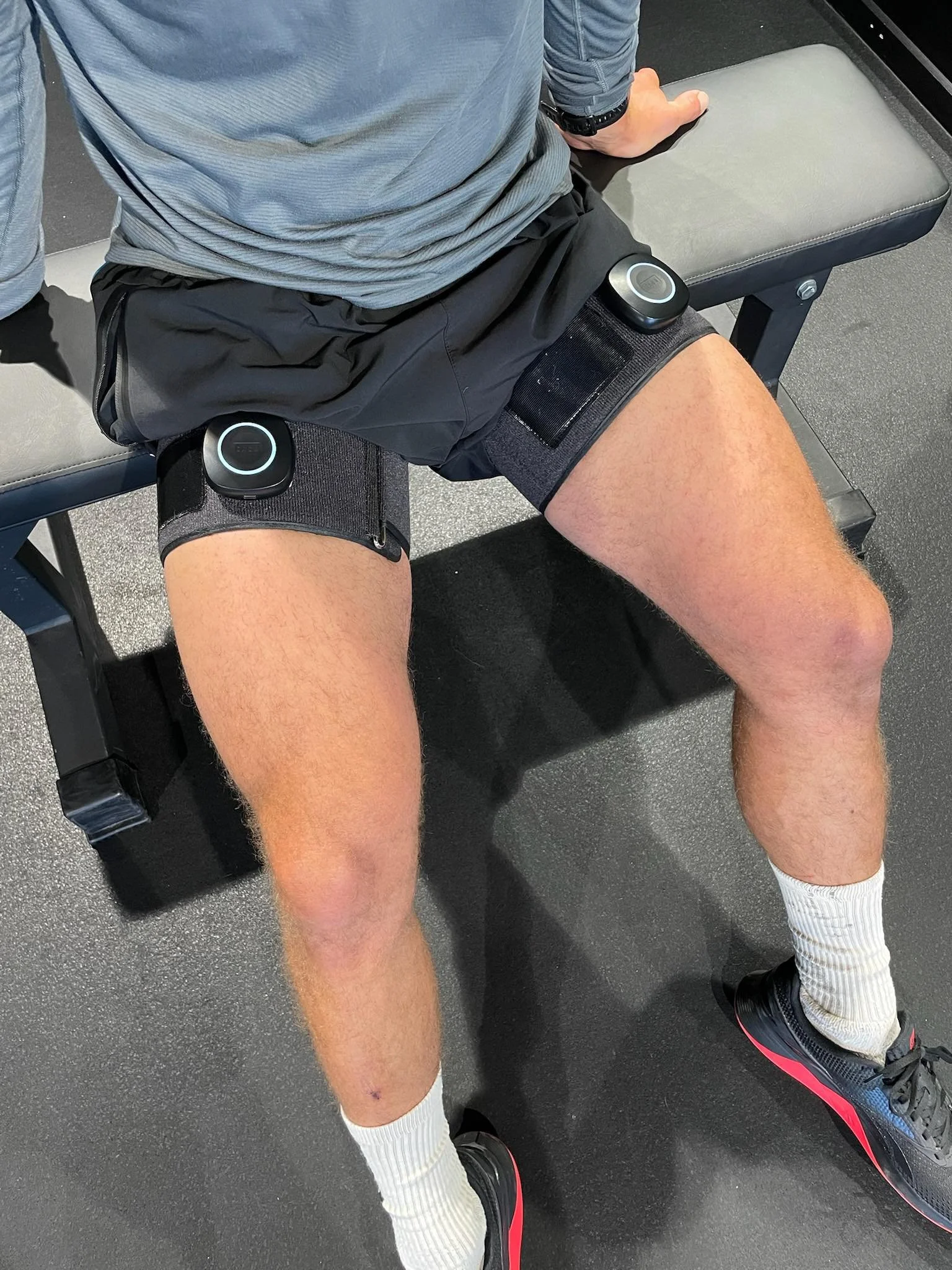 An individual sitting on a gym bench, wearing black shorts with knee straps, white socks, and black athletic shoes with red accents. The person is positioned in a gym setting.