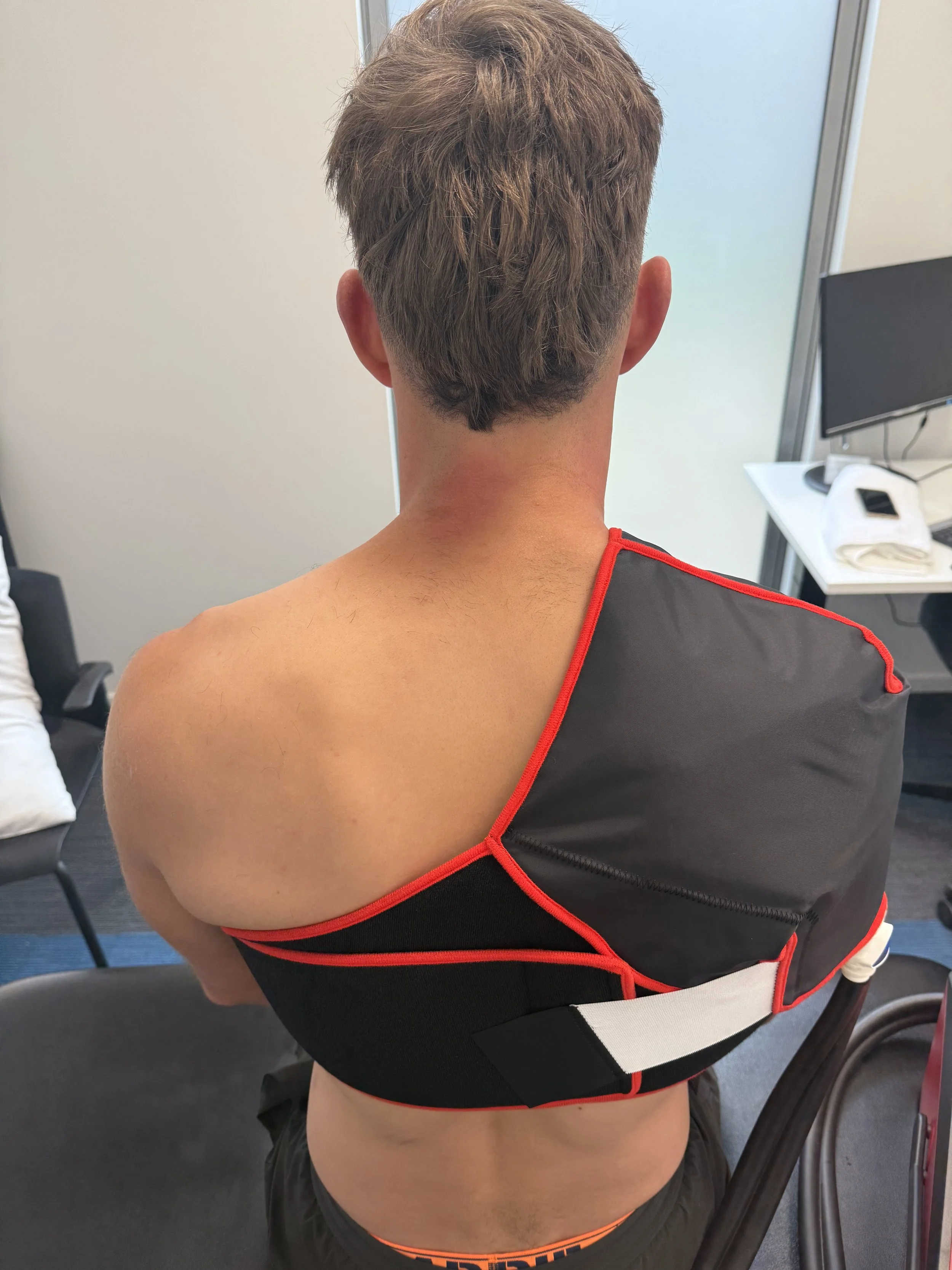 Back view of a shirtless person sitting on a chair, wearing a black shoulder brace with red trim and a white strap, in an office setting with a desk, computer, and chair.