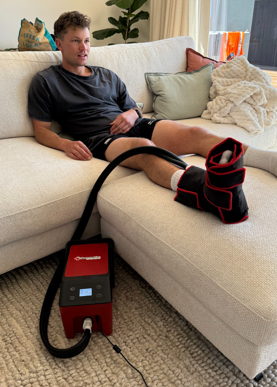 A person sitting on a chair with a leg being treated with a compression therapy device, which is connected to a cryopush cold compression machine fro recovery machine on the floor.