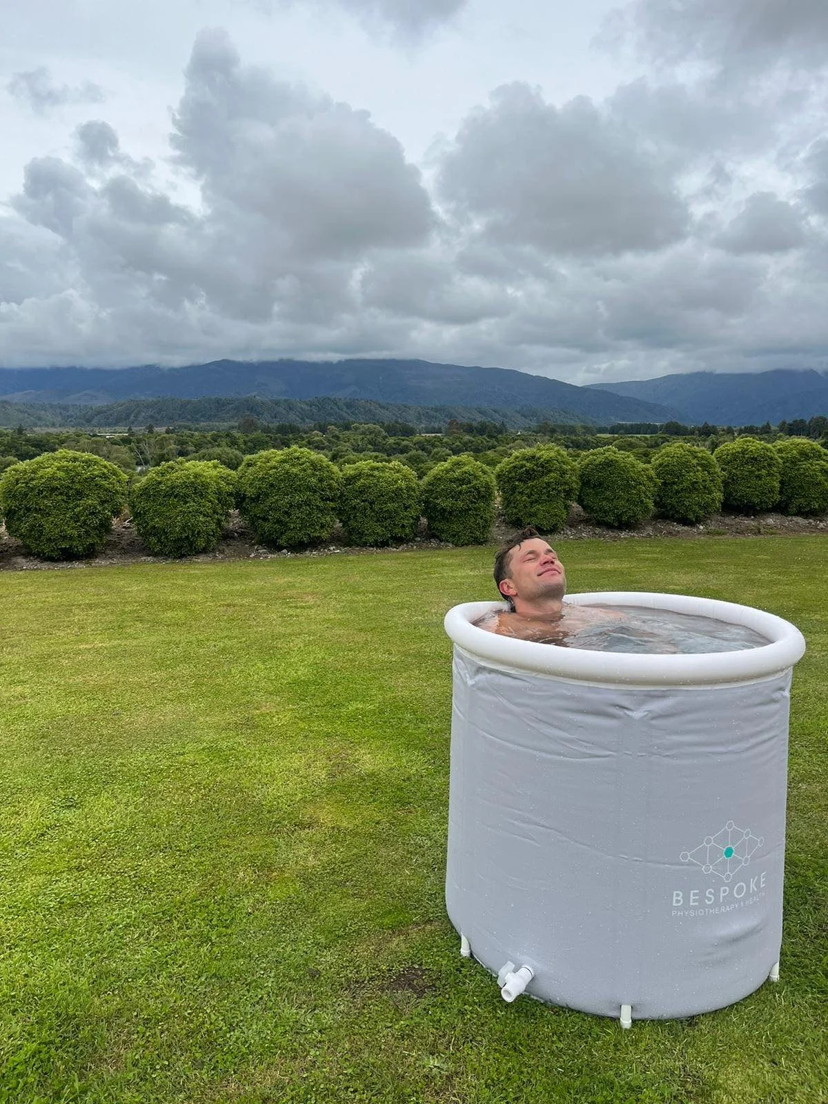 A man relaxing in a portable inflatable ice bath outside on a grassy area, with hills, trees, and cloudy sky in the background.