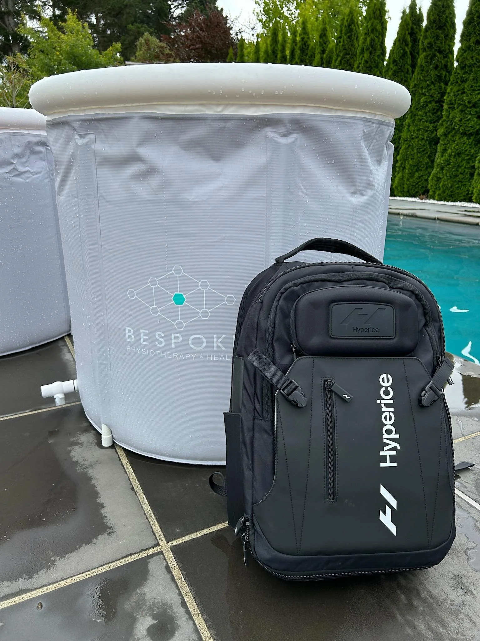 Black Hyperice backpack placed on wet outdoor pool deck next to a large water therapy container with a pool and green hedges in the background.