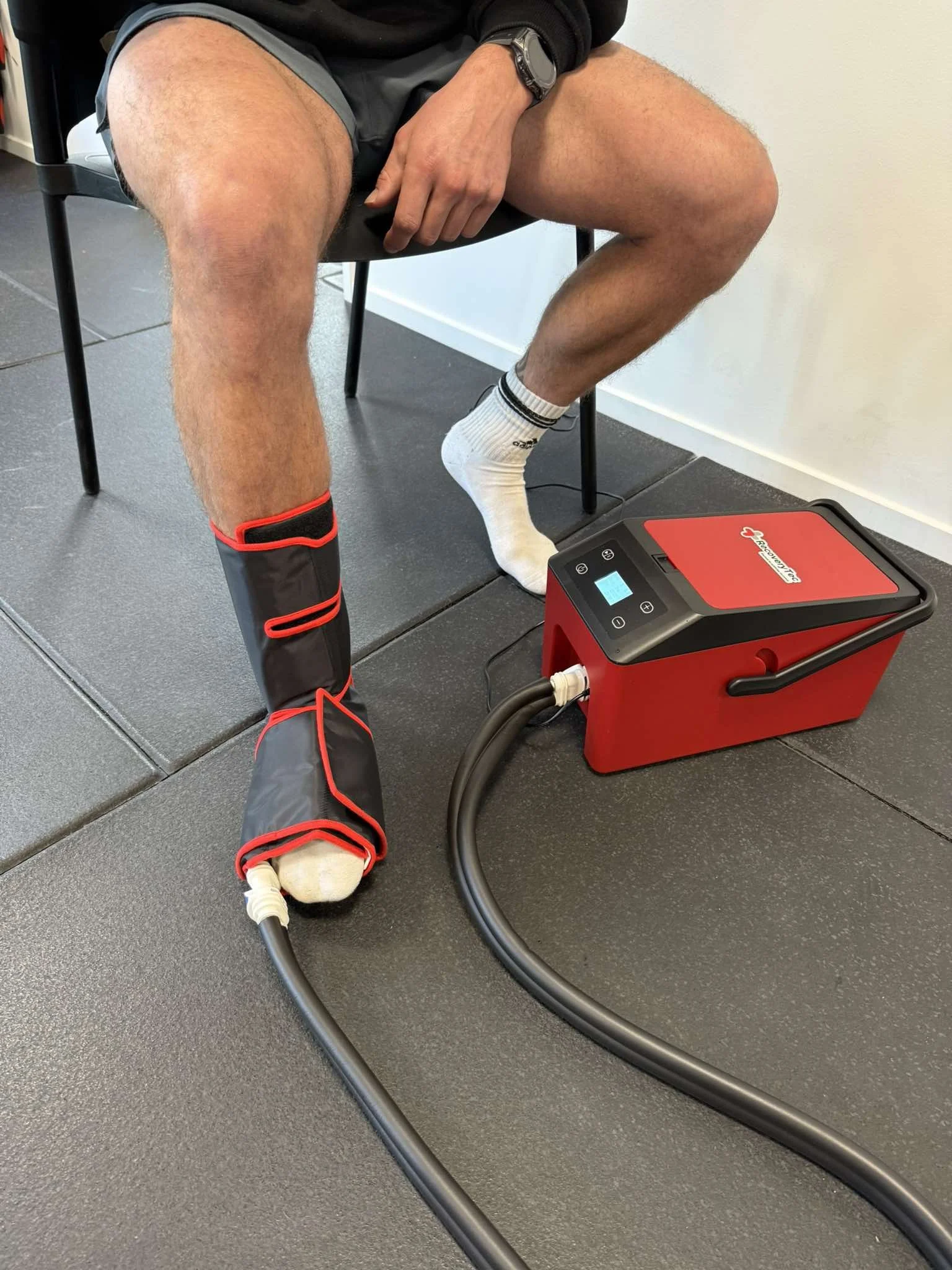 A person sitting on a chair with a leg being treated with a compression therapy device, which is connected to a red machine on the floor.