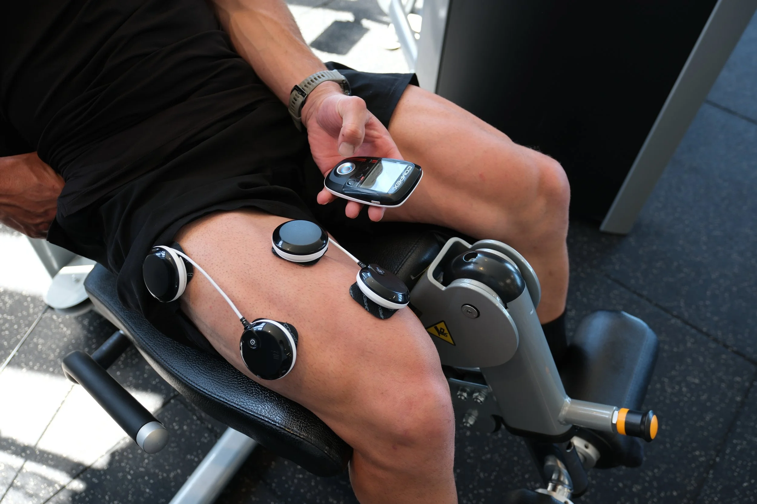 Person sitting with legs extended, holding a device with the brand name 'COMPEX', possibly a muscle stimulator or electronic therapy device.