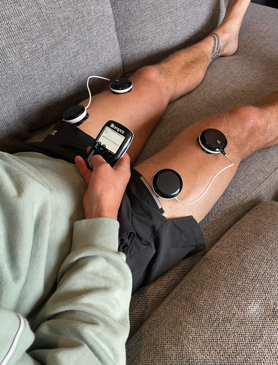 Person sitting with legs extended, holding a device with the brand name 'COMPEX', possibly a muscle stimulator or electronic therapy device.