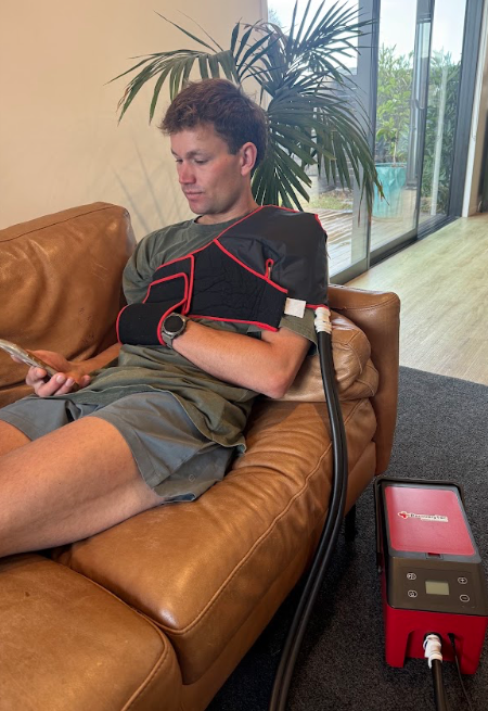 Man wearing a cryopush ice compression machine for recovery from surgery brace on his left shoulder and chest, sitting near a red medical device on a black table.