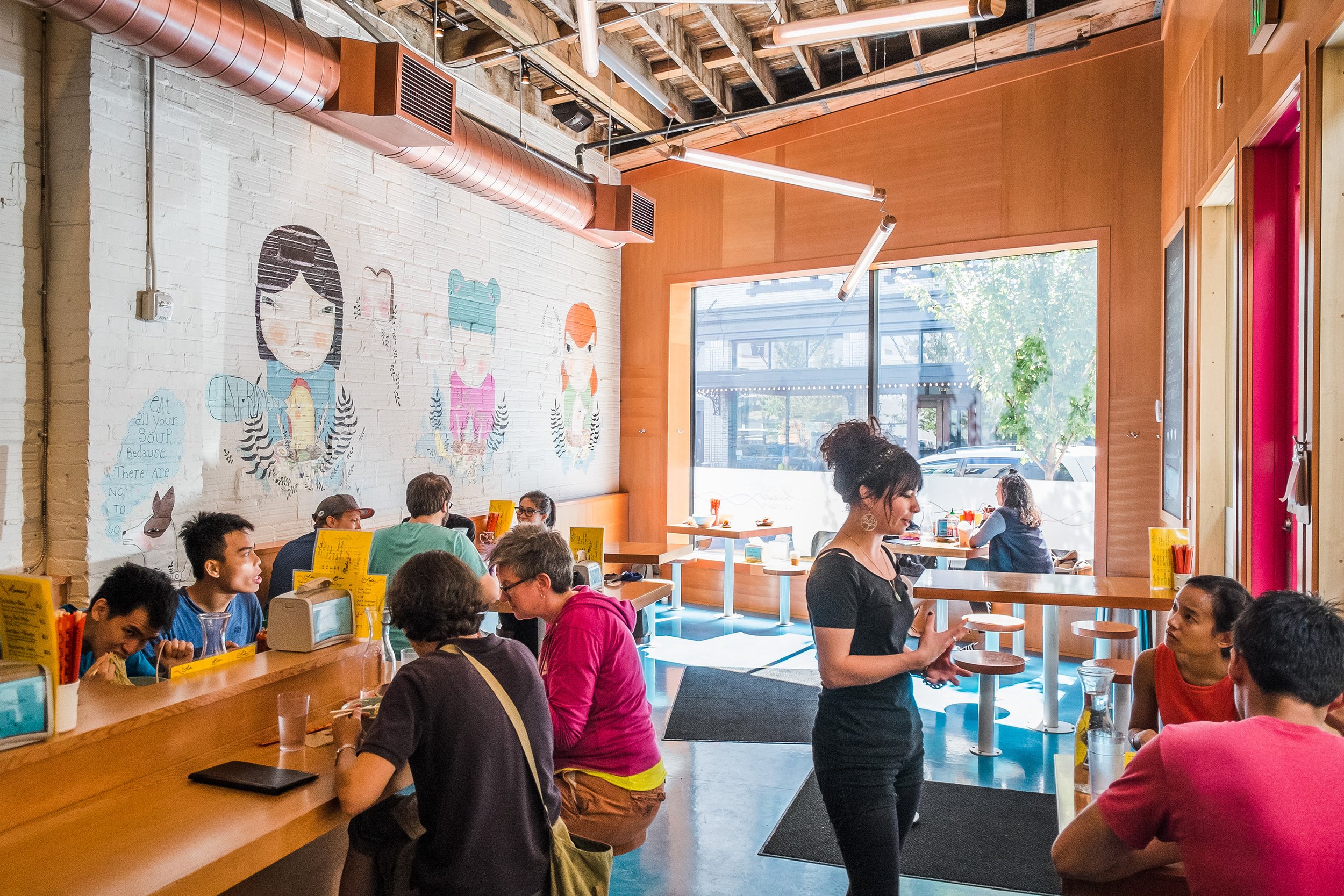 Scene: Busy restaurant with groups of people. Neutral, warm colors dominate—white and brown. Foreground: Woman in black dress conversing with a man over a meal. Background: Vibrant pink-hooded woman, laughing man with beard, diverse crowd. Blurred wi