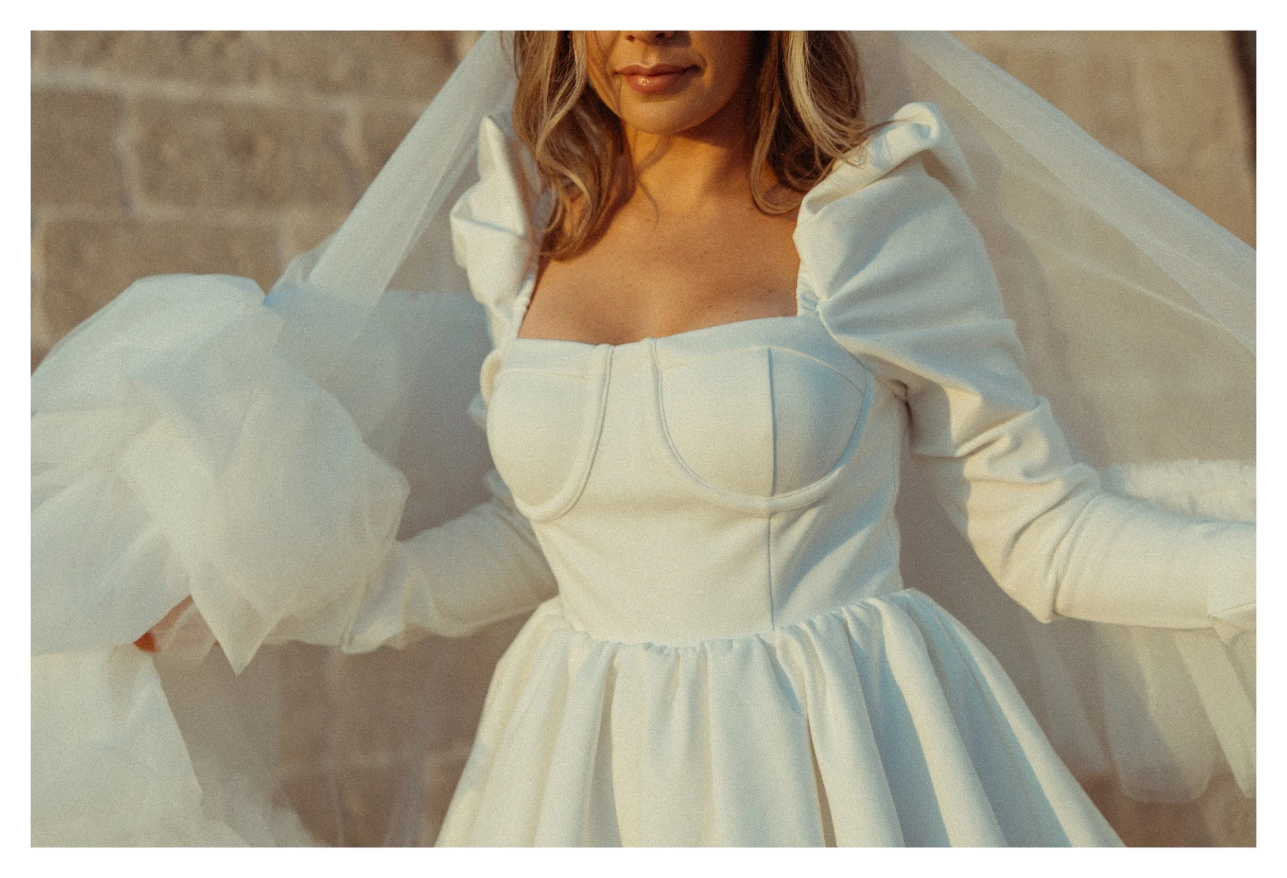A woman in a white dress with puffed sleeves and a fitted bodice, standing outdoors against a stone wall, with sunlight illuminating her face and dress.