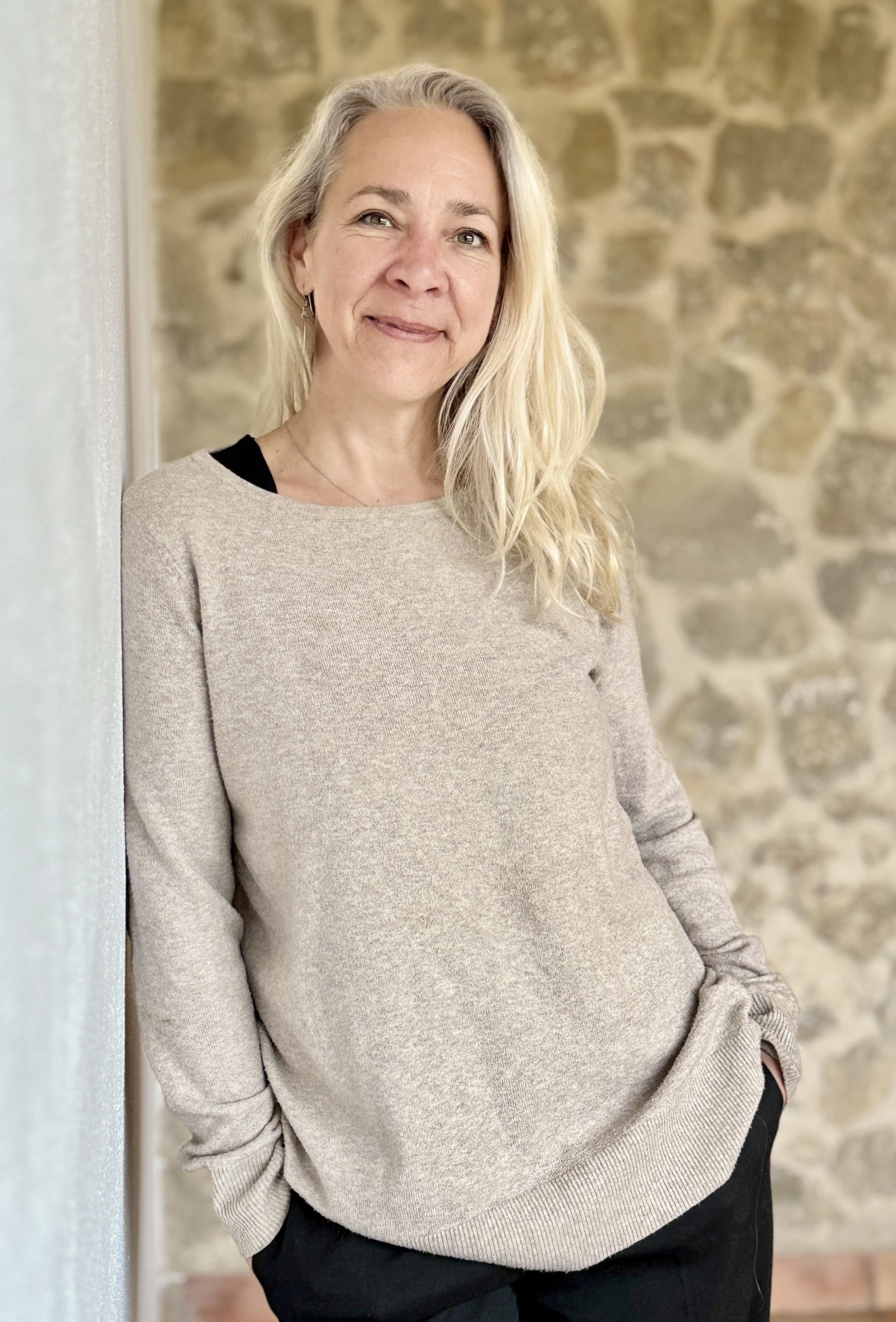 A woman with long blonde hair, wearing a beige sweater and black pants, leaning against a wall with a brick background and smiling at the camera.