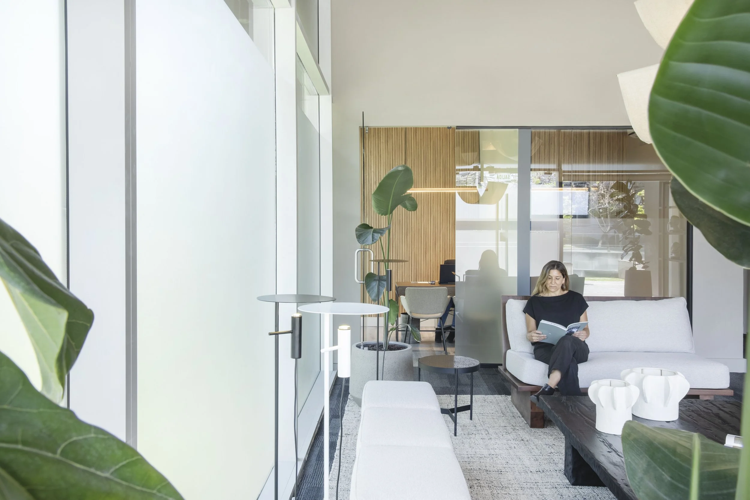 Woman reading a book on a white sofa in a modern, plant-filled office lounge with glass walls and wood paneling