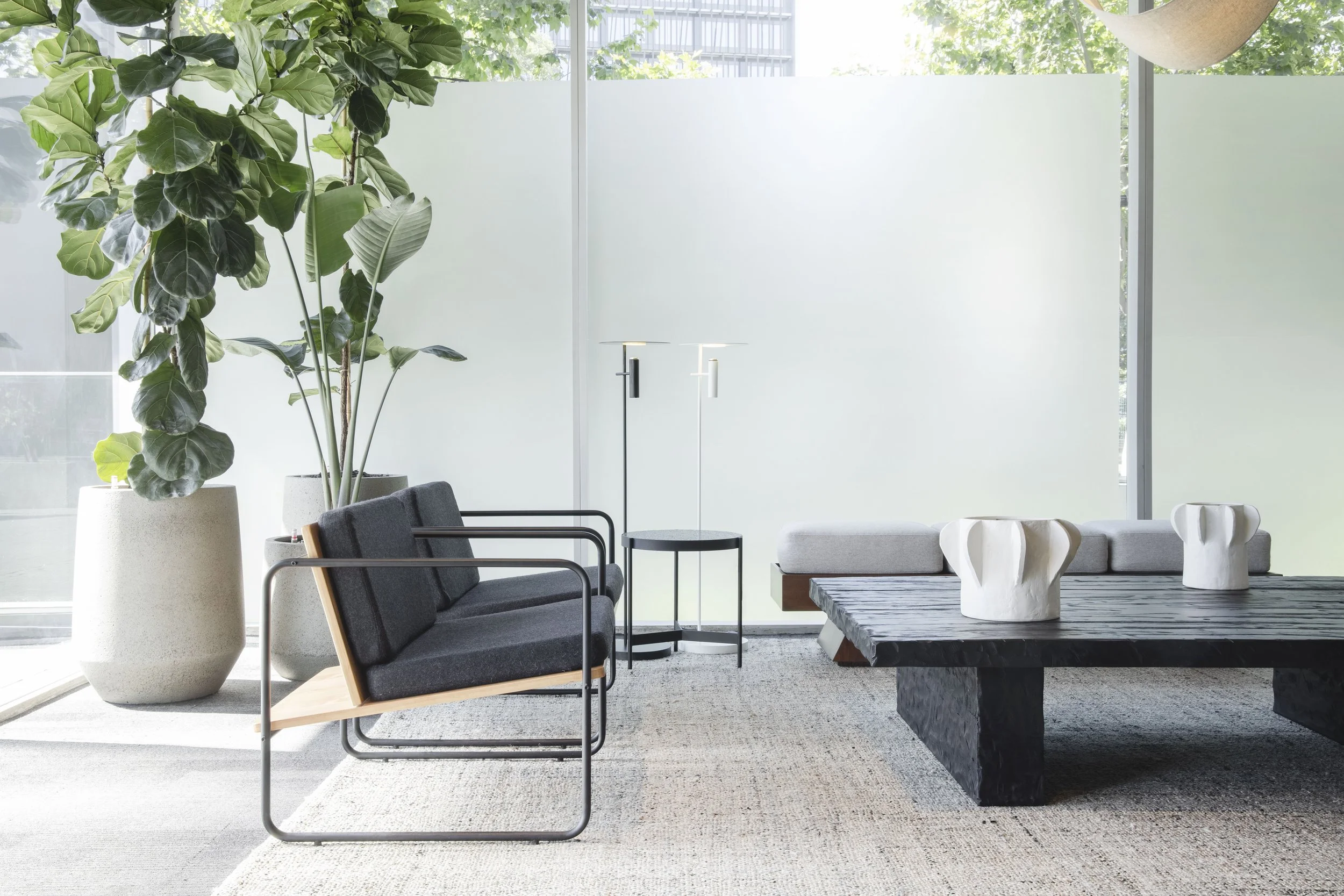 Bright modern living room with large potted plants, gray lounge chairs, sofa, black coffee table, and sculptural white vases