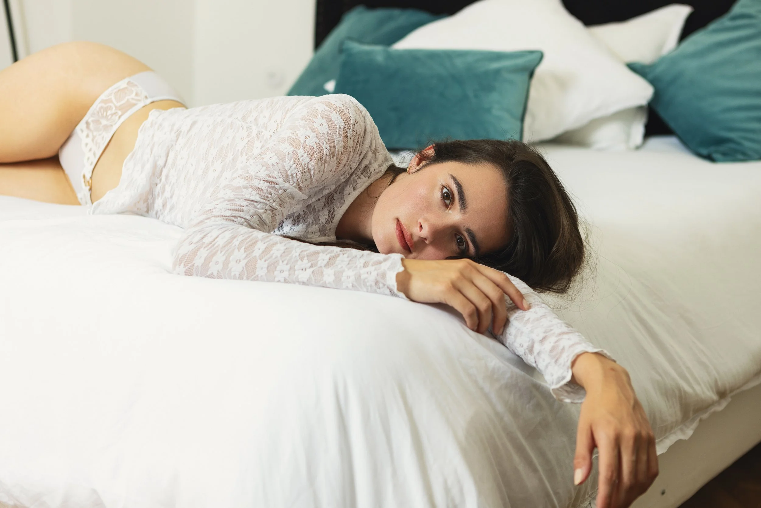 Woman in white lace lingerie lying across a bed in a softly lit intimate interior.