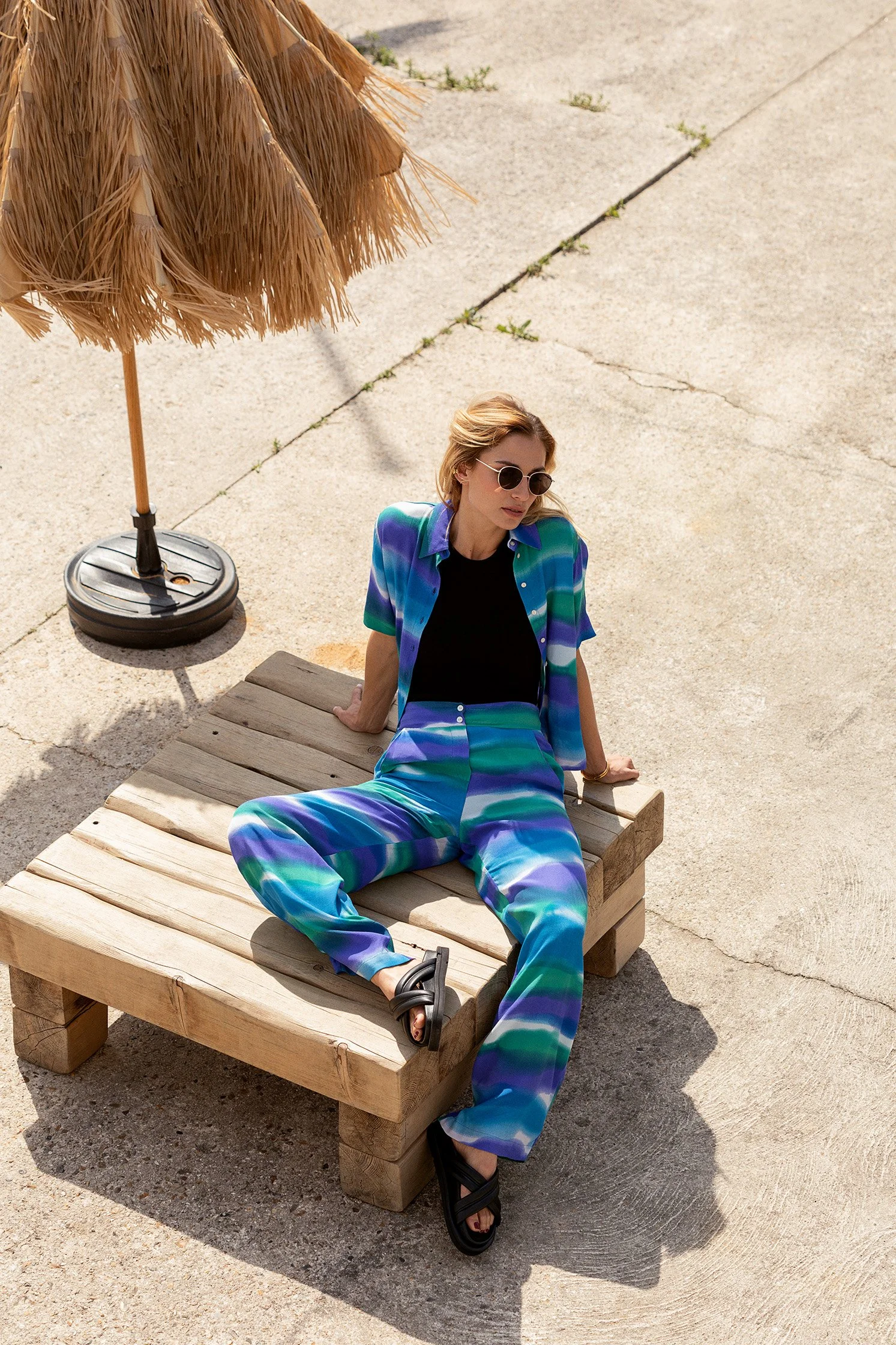 Overhead fashion photograph of a woman in a blue and green outfit seated outdoors in strong sunlight.