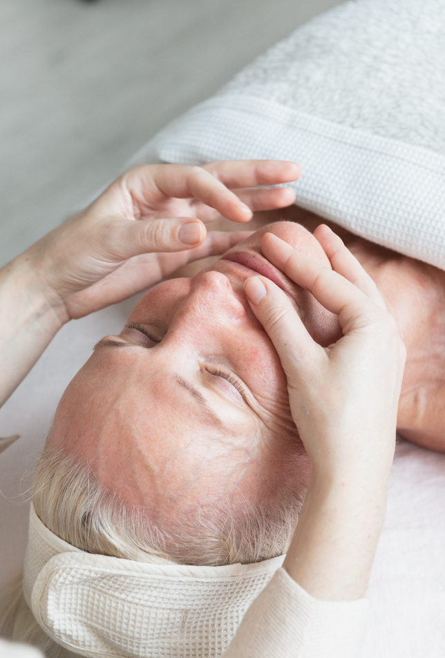 Close-up of an older woman receiving a gentle natural facial massage with her eyes closed.