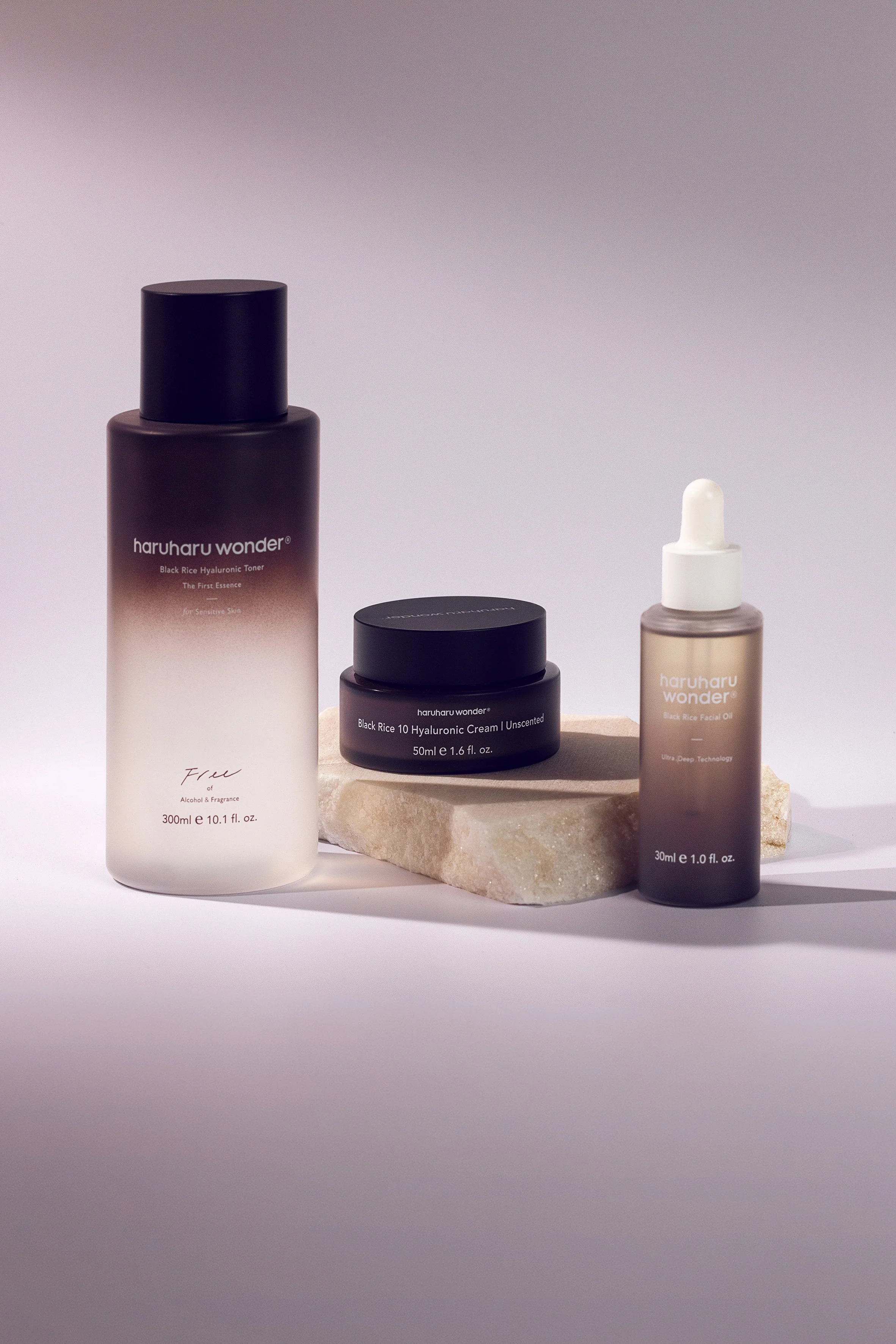 Three Haruharu Wonder skincare products arranged on a stone pedestal in soft studio light against a muted mauve background.