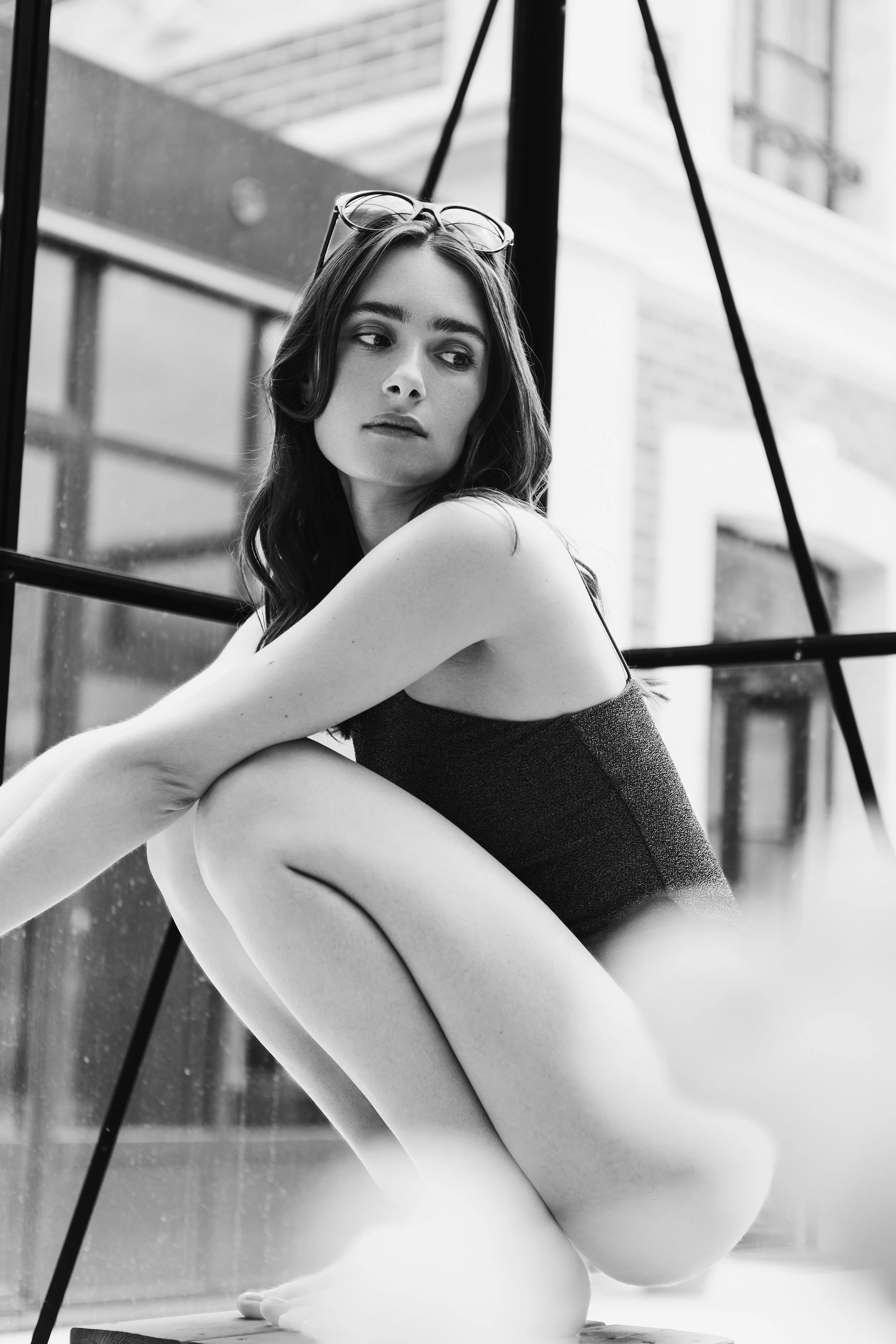 Black and white editorial photograph of a seated woman in a dark bodysuit beside large windows.