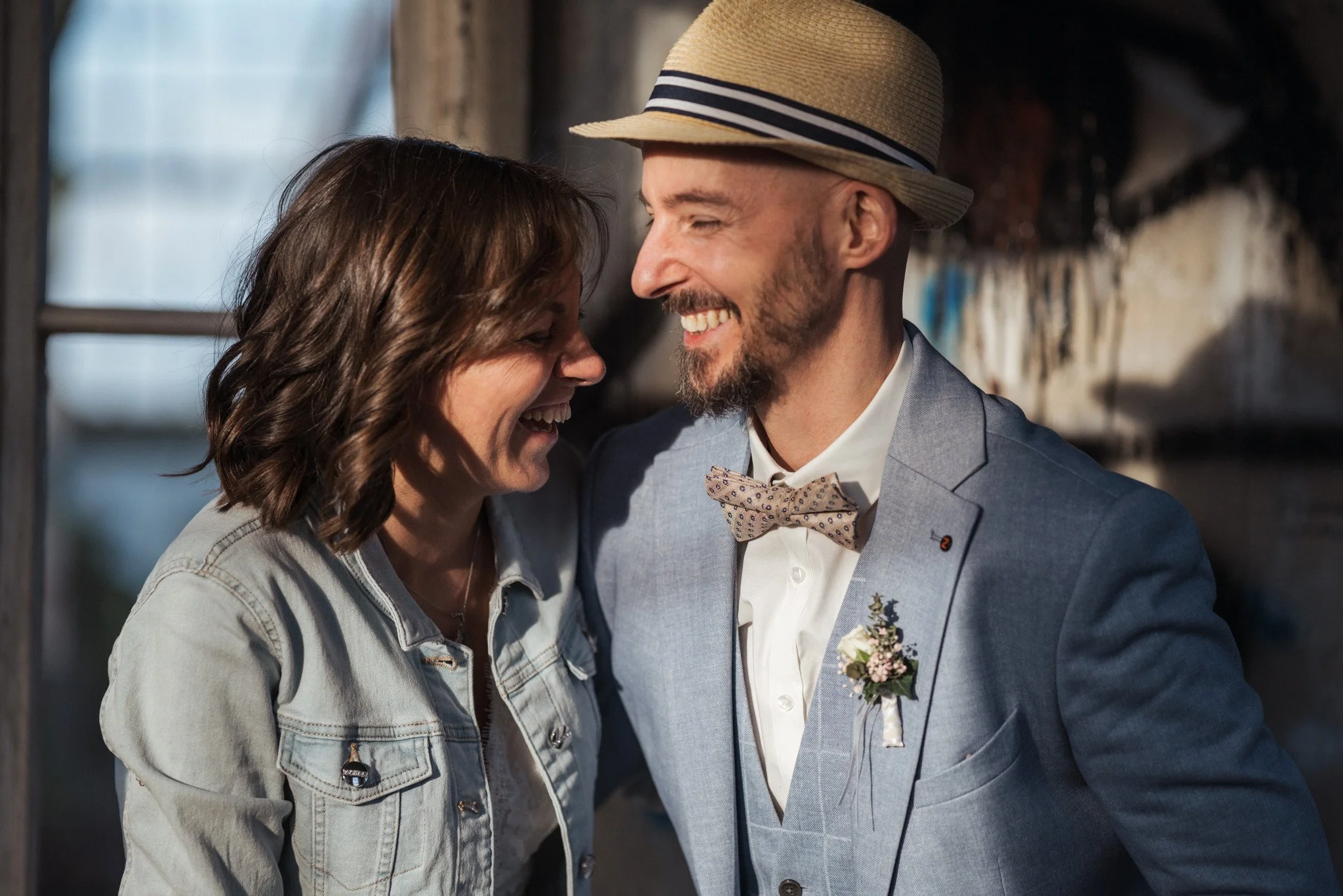 Ein lachendes Paar bei einer Hochzeit, der Mann trägt einen Anzug mit Fliege und einen Hut, die Frau trägt eine Jeansjacke, sie scheinen eine glückliche Moment zu teilen.