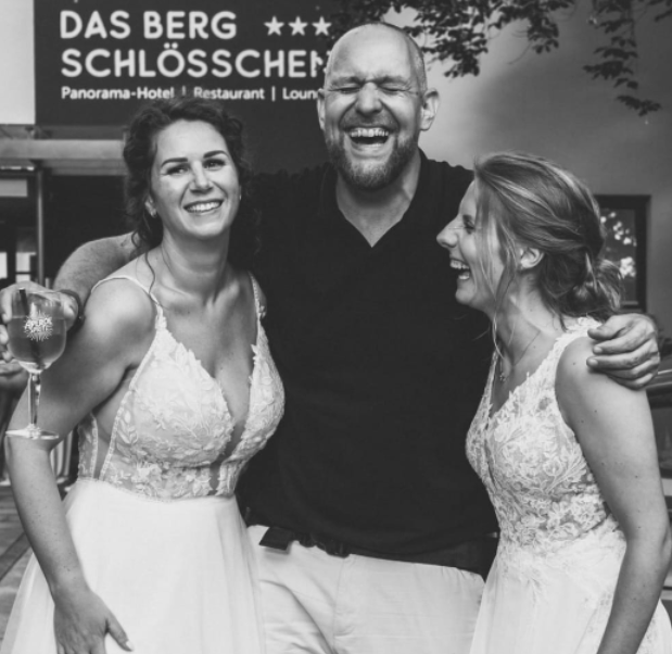 Drei Menschen lachen und umarmen sich, zwei Frauen in weißen Kleidern, ein Mann in schwarzem T-Shirt, im Hintergrund ein Schild mit dem Text "Das Berg Schlösser".