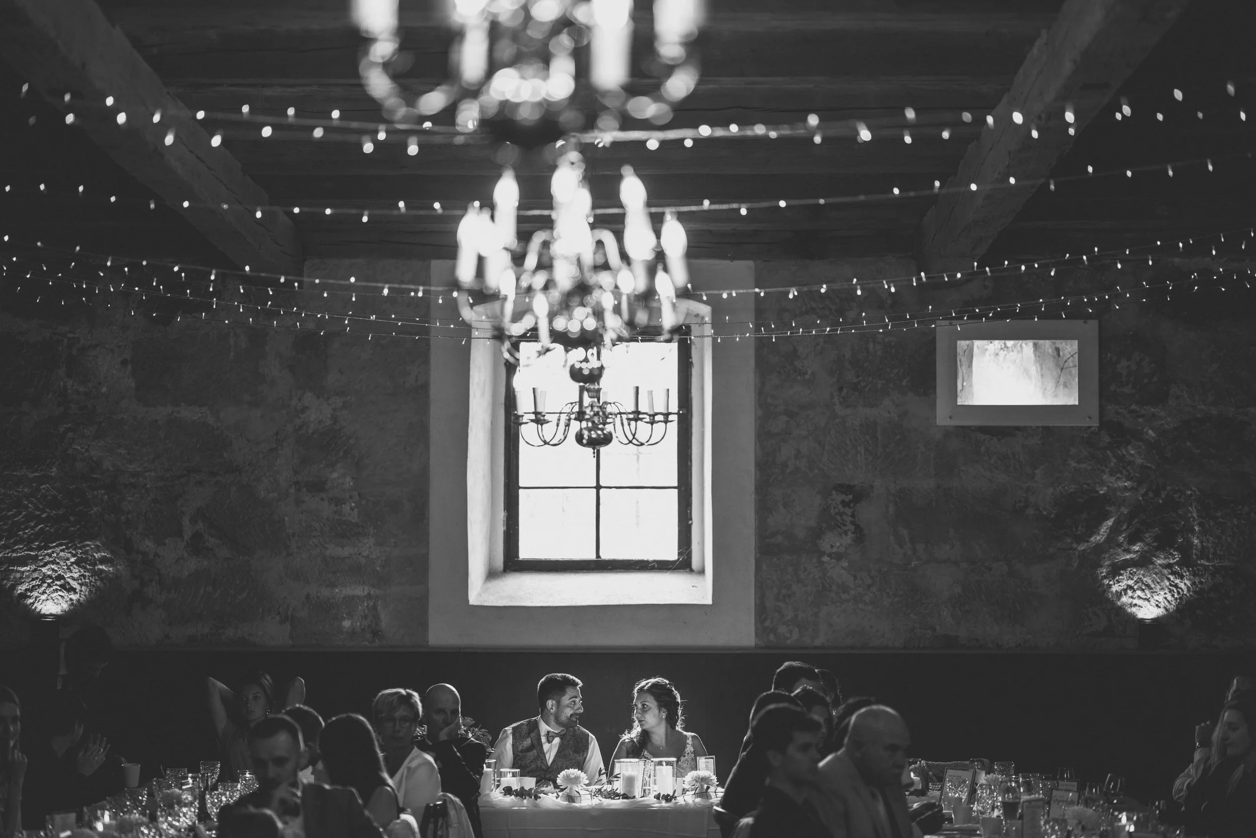 Gäste sitzen an einem festlich gedeckten Tisch in einer rustikalen, dunkel verputzten Scheune, beleuchtet von Lichterketten und einem Kronleuchter, während im Hintergrund ein Paar spricht.