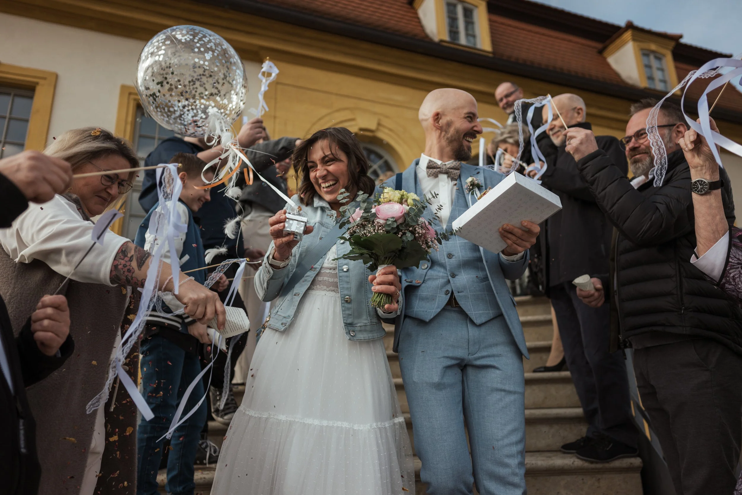 Ein frisch verheiratetes Paar, ein Mann und eine Frau, steht auf Treppen vor einem historischen Gebäude. Die Frau trägt ein weißes Hochzeitskleid und hält einen Blumenstrauß, während der Mann einen Geschenkkarton hält. Sie sind von Freunden und Famil