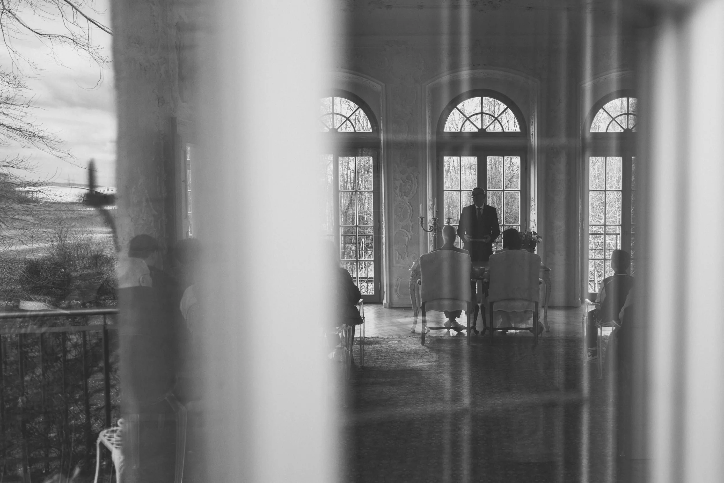 Ein schwarz-weiß-Bild zeigt eine elegante Innenraumaufnahme eines Raumes mit großen, hohen Fenstern und einem Mann in einem Anzug, der sich wahrscheinlich auf einer Veranstaltung oder in einer Zeremonie befindet. Der Blick ist durch eine halboffene Tür oder einen Vorhang verzerrt.