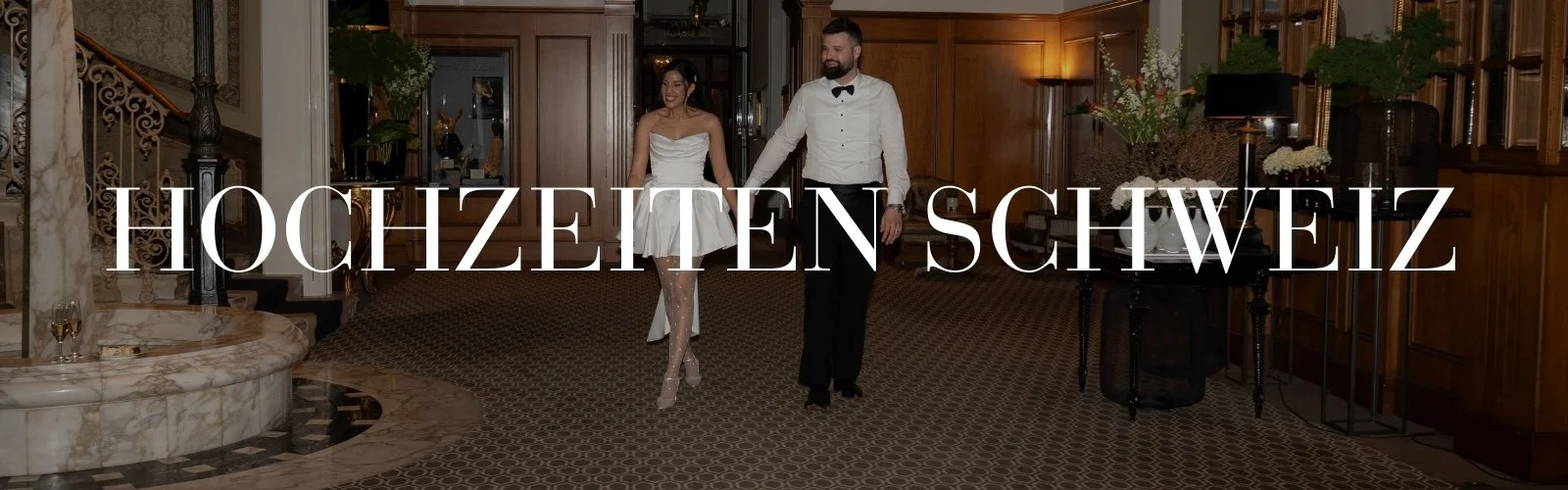 Ein Brautpaar tanzt in einem eleganten Hotel-Lobby, die Frau trägt ein weißes Brautkleid und der Mann einen weißen Hemd und schwarzen Anzug. Der Raum ist mit Holzvertäfelung, Blumen und Kerzen dekoriert, mit dem Text "HOCHZEITEN SCHWEIZ" überlagert.