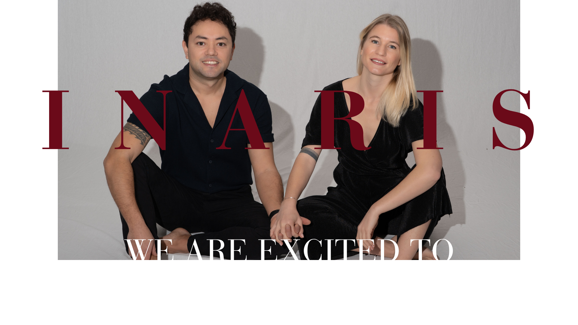 A man and a woman are sitting on the floor, holding hands. The woman is wearing a black dress, the man a black shirt. The background is a white wall.